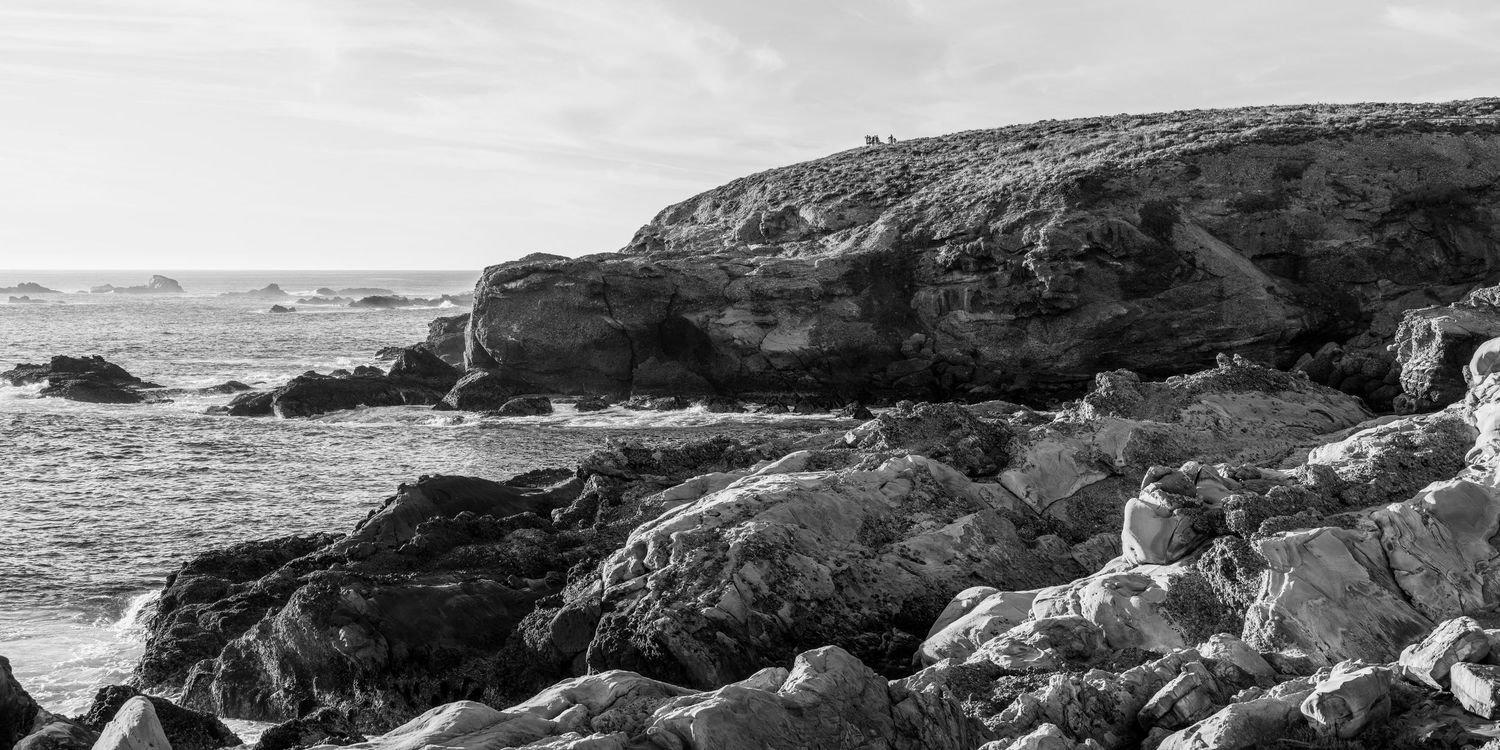 Black and white seascape with rugged rocky coastline and gentle ocean waves under a clear sky.