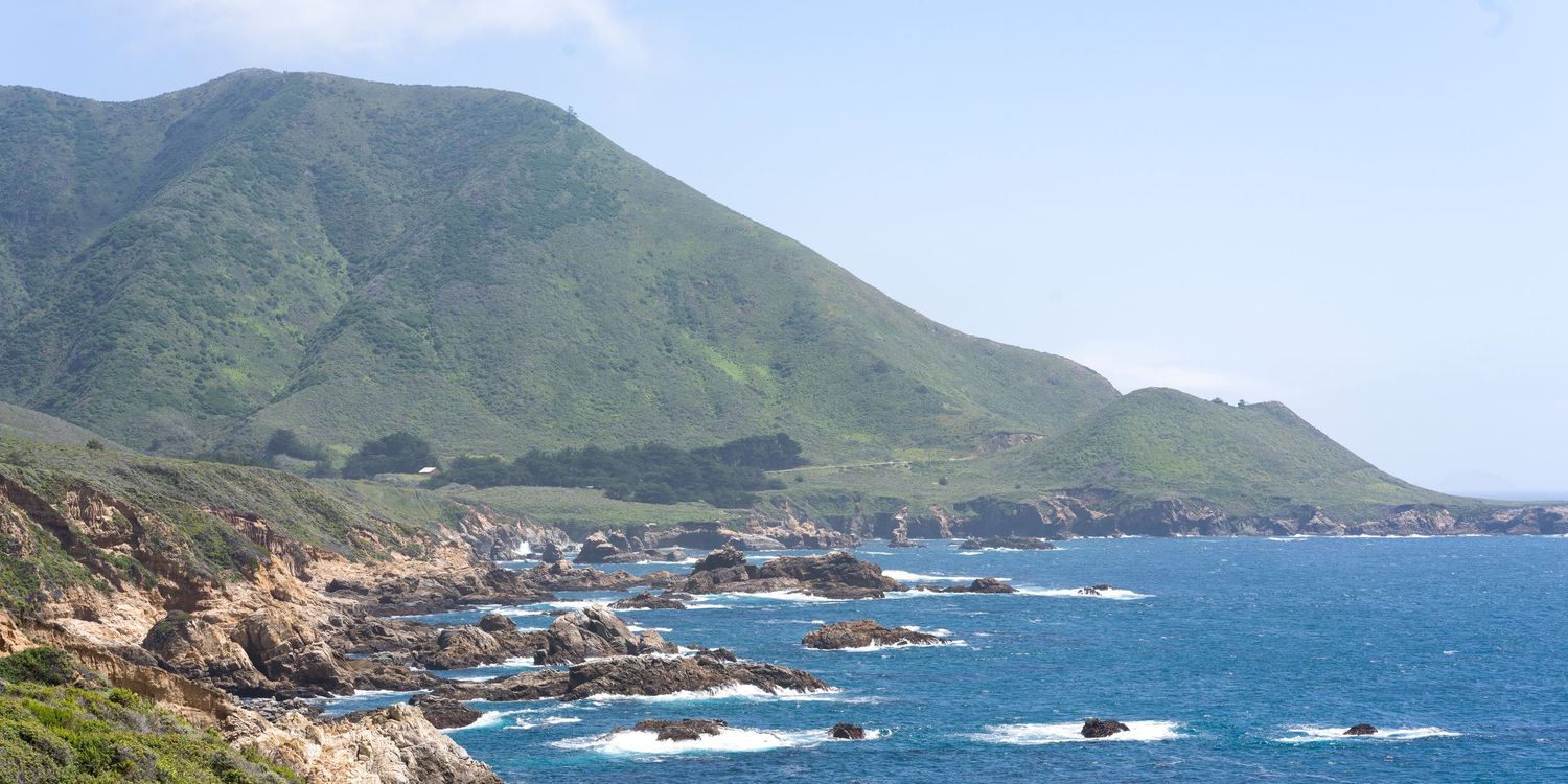 Lush green mountain slopes meet rocky Pacific coastline in Big Sur with vivid blue ocean waters under clear skies.