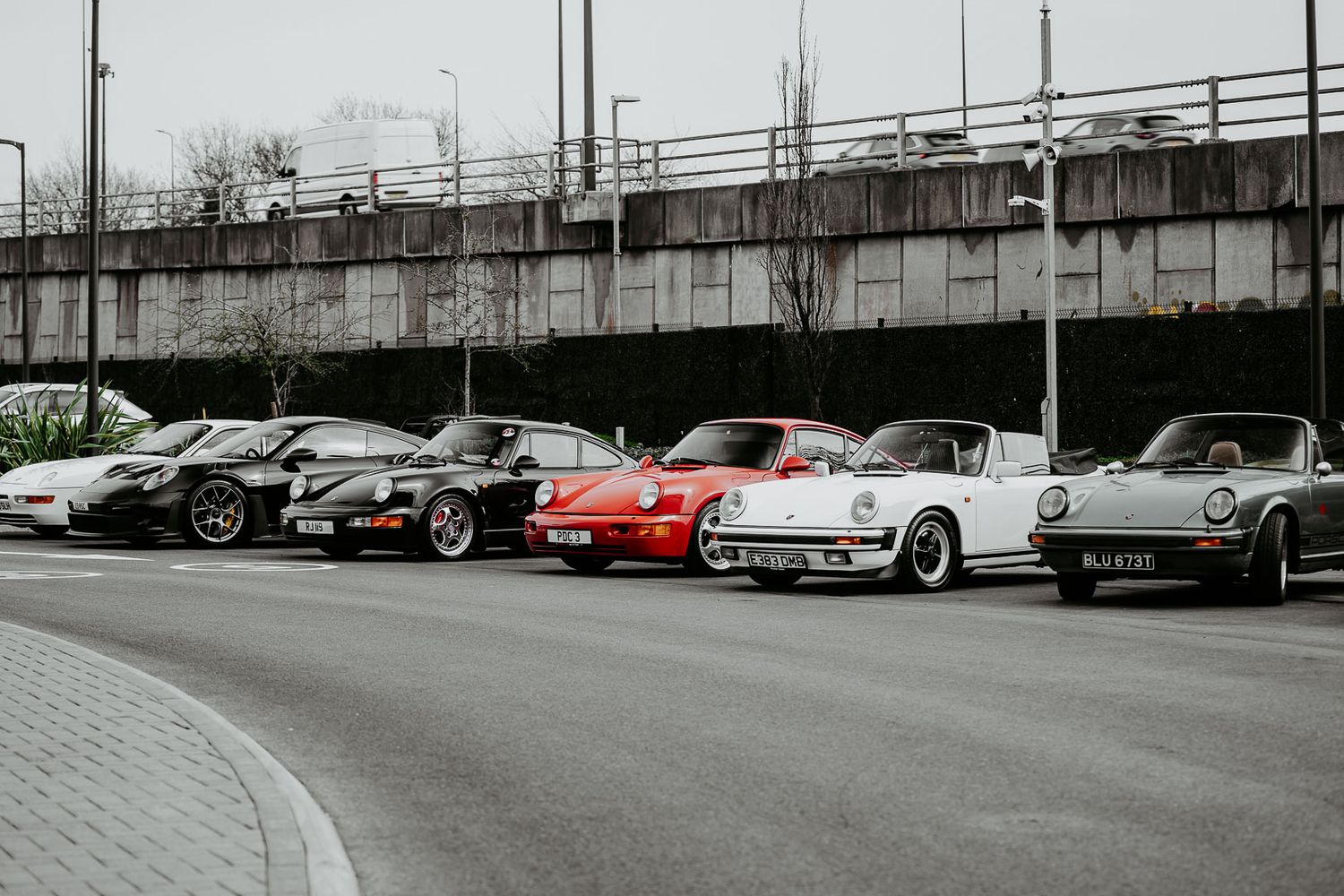 Four classic Porsche 911 models lined up on a road in front of a concrete bridge overpass.