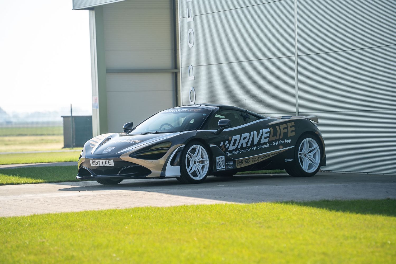 McLaren 720S with DriveLife livery wrap parked outside an aviation hangar on a sunny airfield.