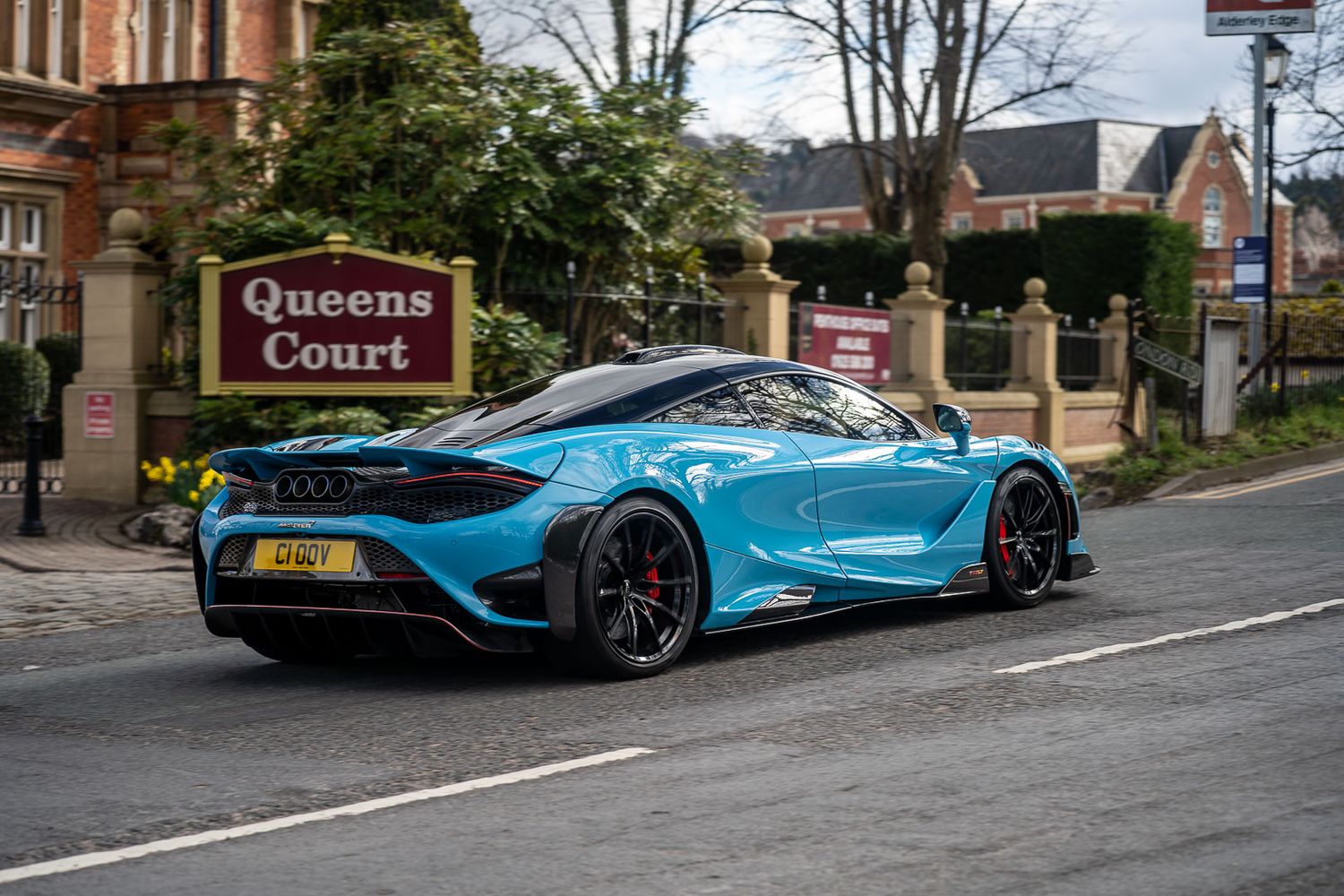 Bright blue McLaren 720S supercar driving past Queens Court sign on a tree-lined suburban street.