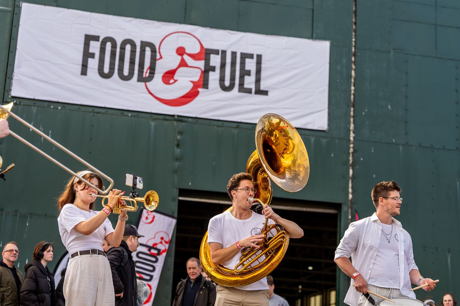 Brass band musicians performing outside a Food and Fuel branded industrial building.