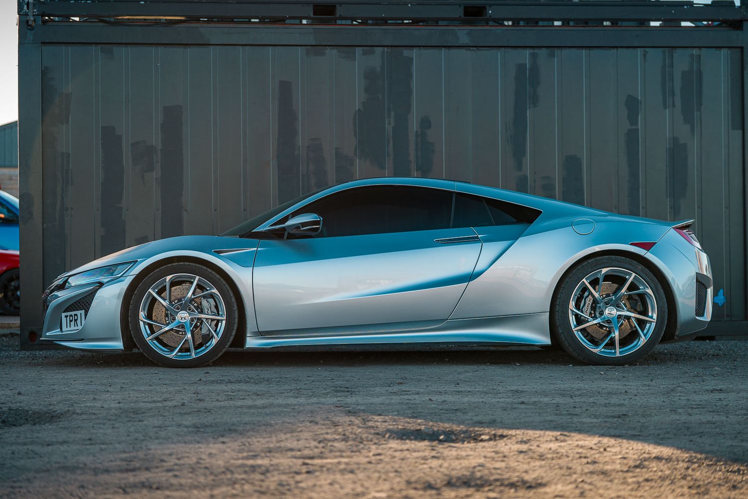 Silver Honda NSX supercar parked in profile view against an industrial metal backdrop.