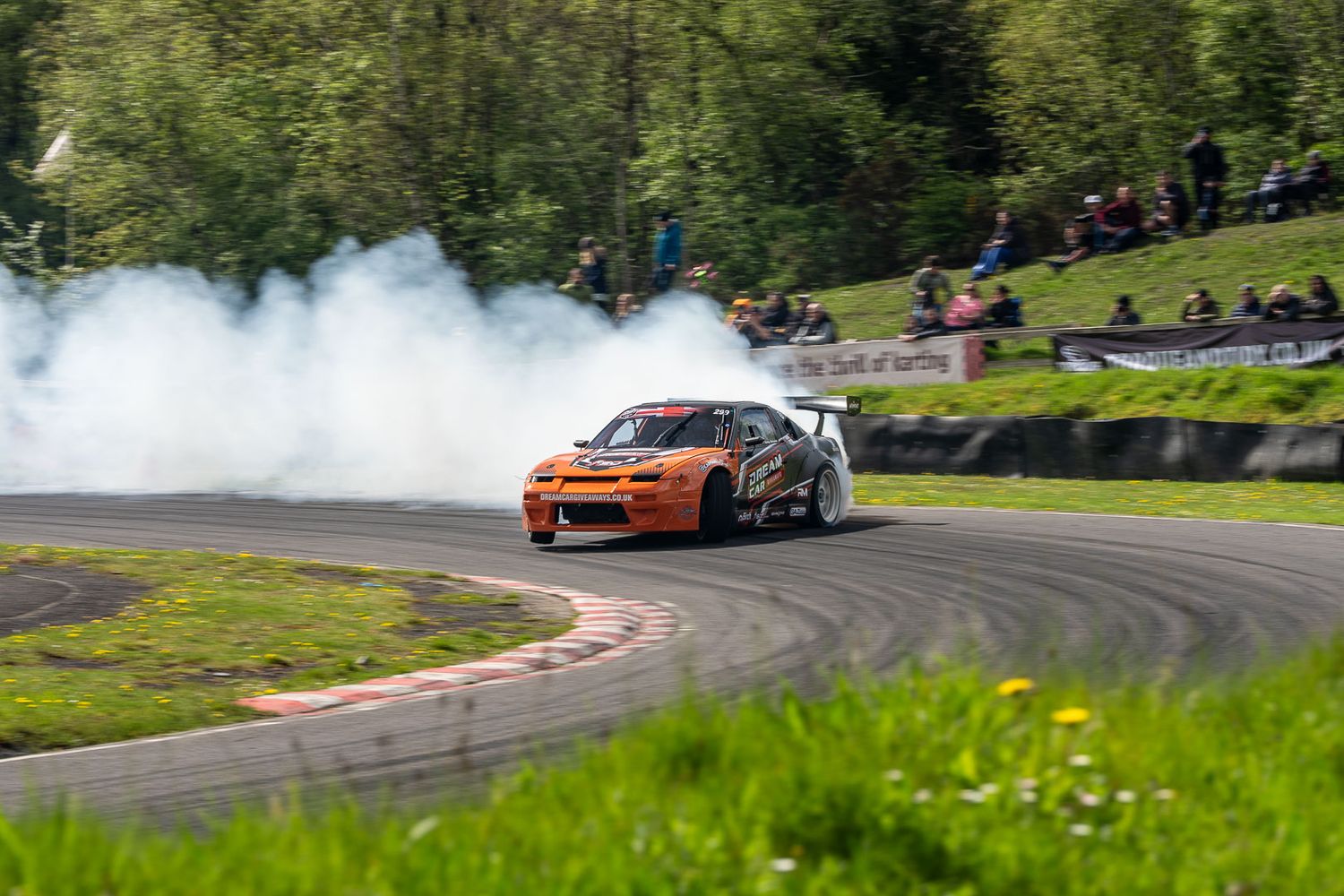 Orange drift car producing smoke while cornering on a race circuit with spectators.