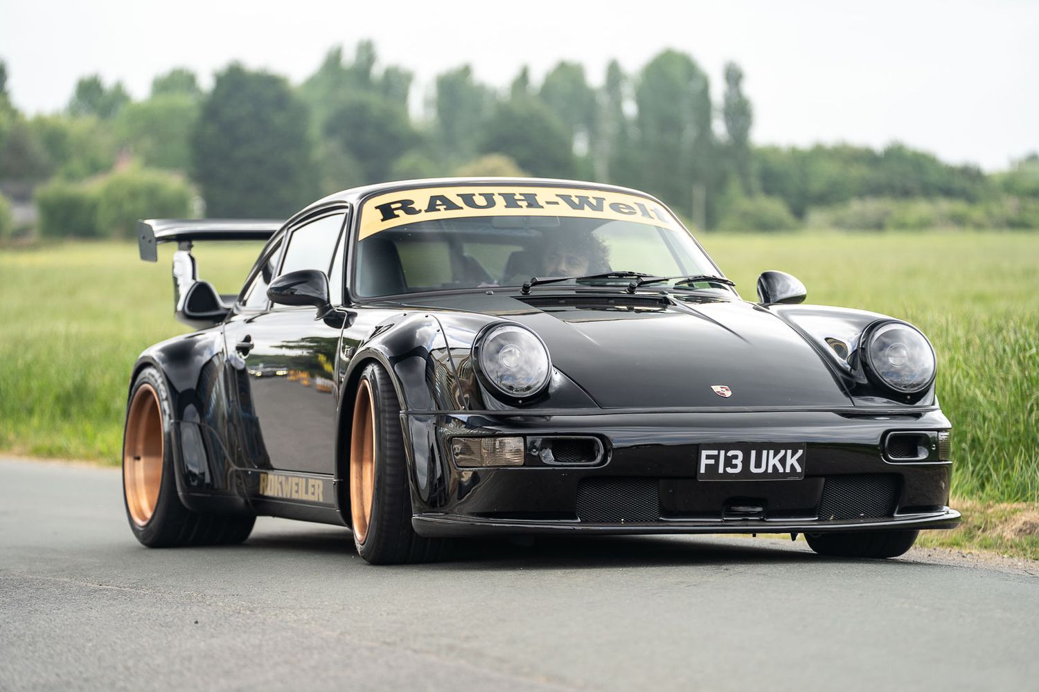 Black RWB Porsche 911 with wide body kit and gold wheels on a country road.