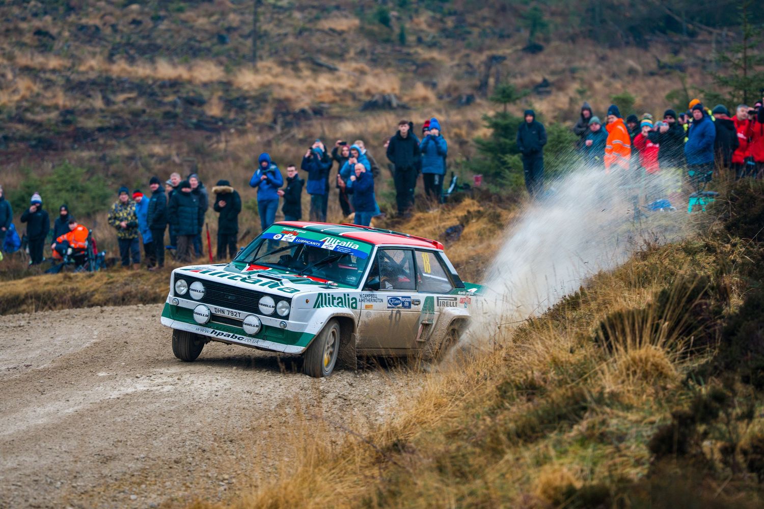 Classic Fiat 131 Abarth rally car sliding on a gravel stage with spectators watching from the hillside.