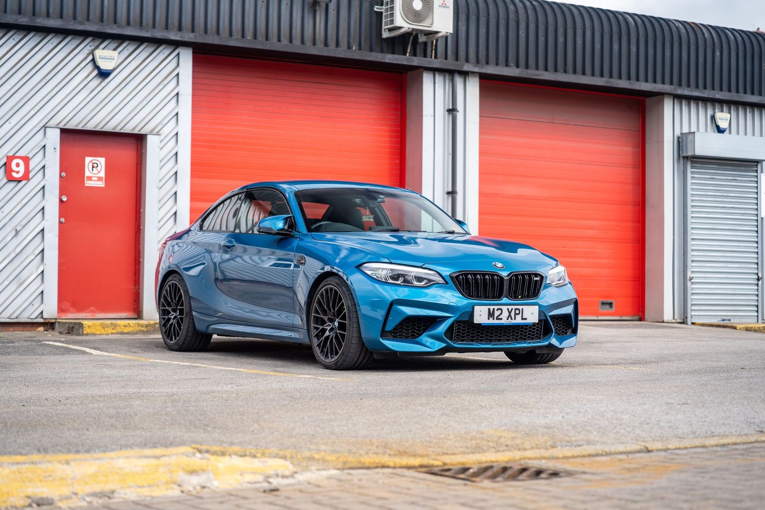 Blue BMW M2 Competition parked in front of red roller shutter doors at an industrial unit.