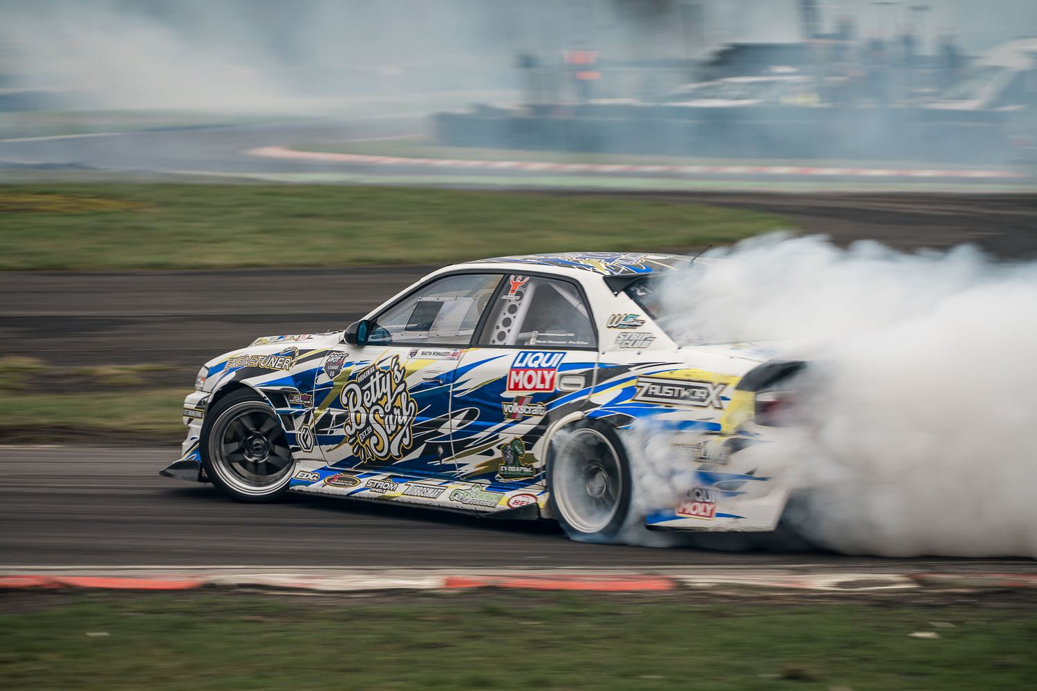 Rally car drifting with massive smoke clouds on a dirt track during a motorsport competition event.