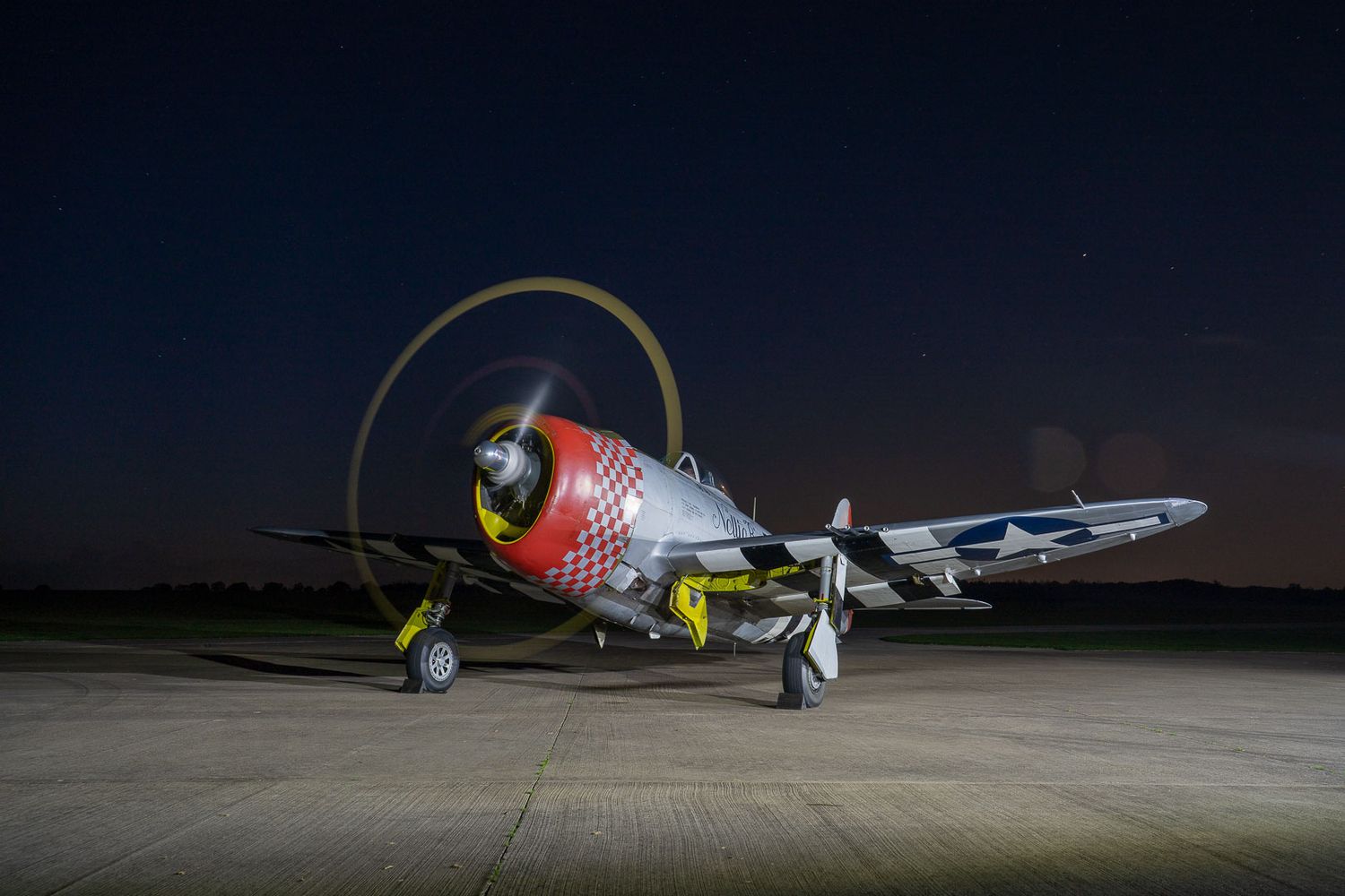 Vintage WWII P-47 Thunderbolt fighter aircraft illuminated on a runway under a night sky.