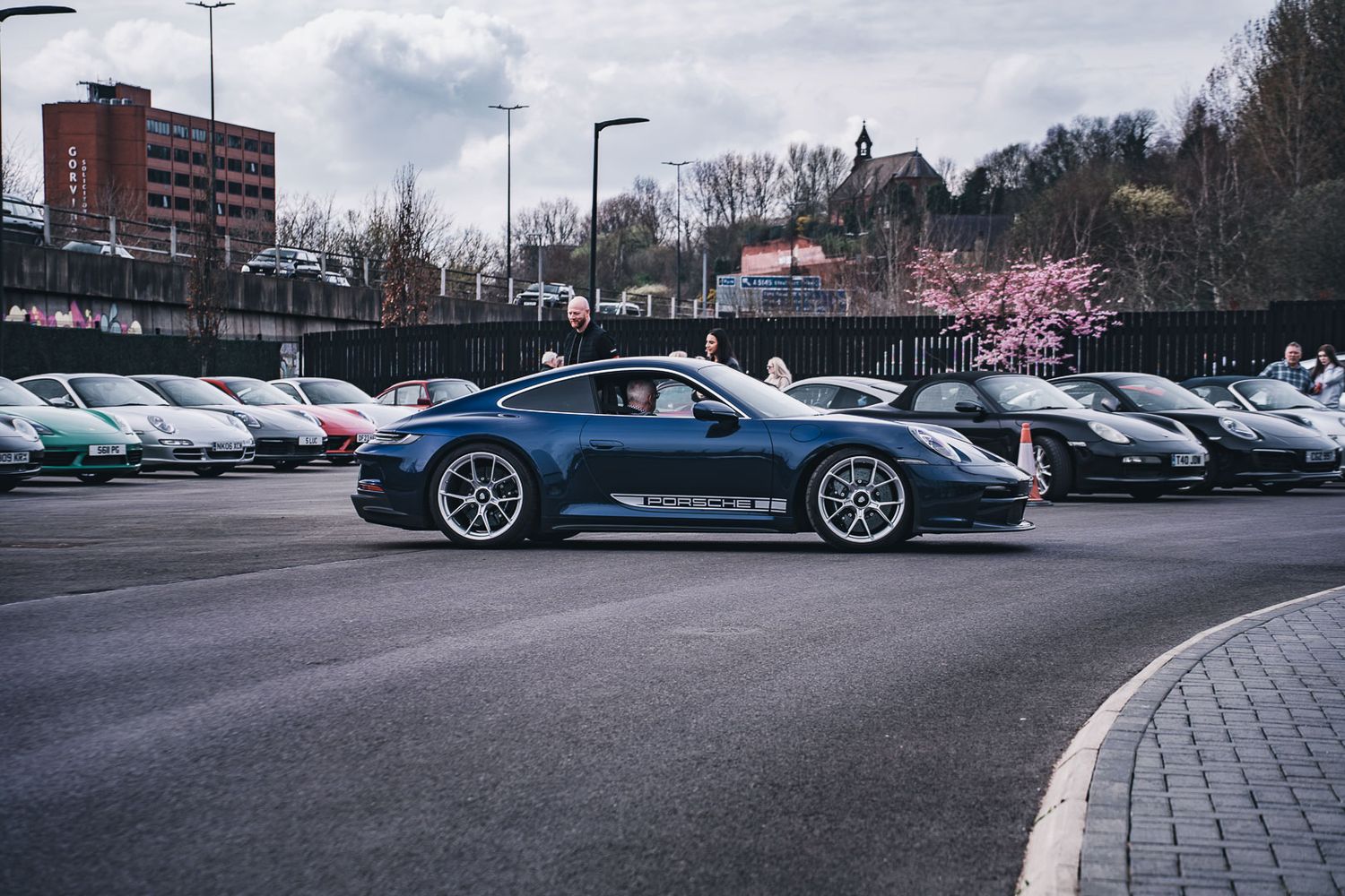 Dark blue Porsche 911 GT3 parked among other Porsches in an urban car park with cherry blossom trees.