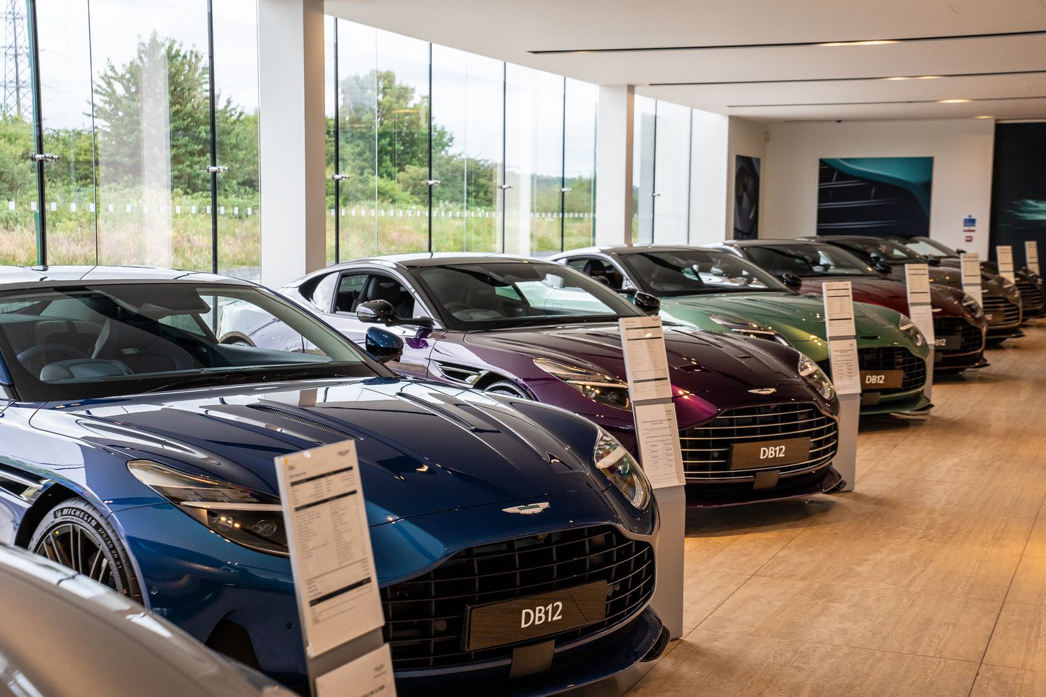 Row of Aston Martin DB12 luxury sports cars in various colors displayed in a modern glass dealership showroom.