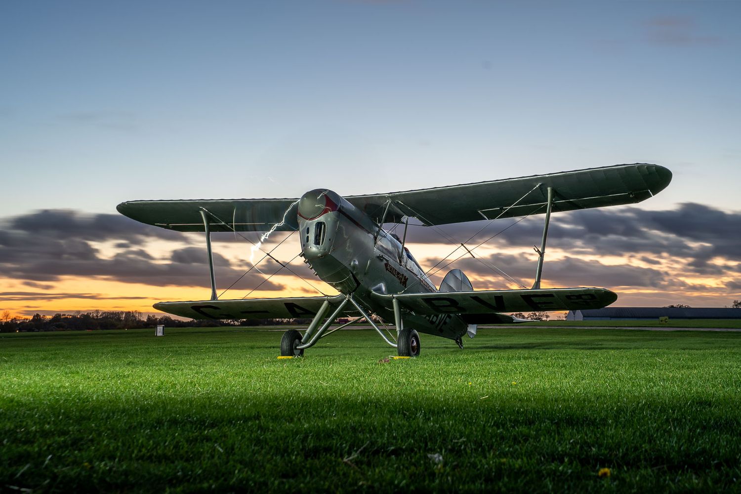Vintage biplane parked on green grass airfield at sunset with dramatic colorful clouds in the background.