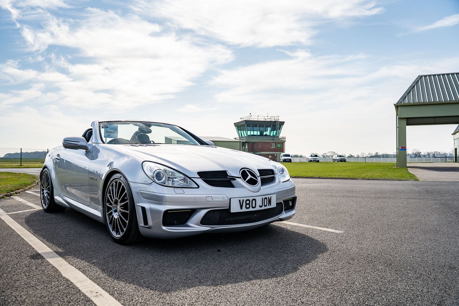 Silver Mercedes-Benz SLK AMG convertible parked on tarmac near an airfield control tower under blue skies.