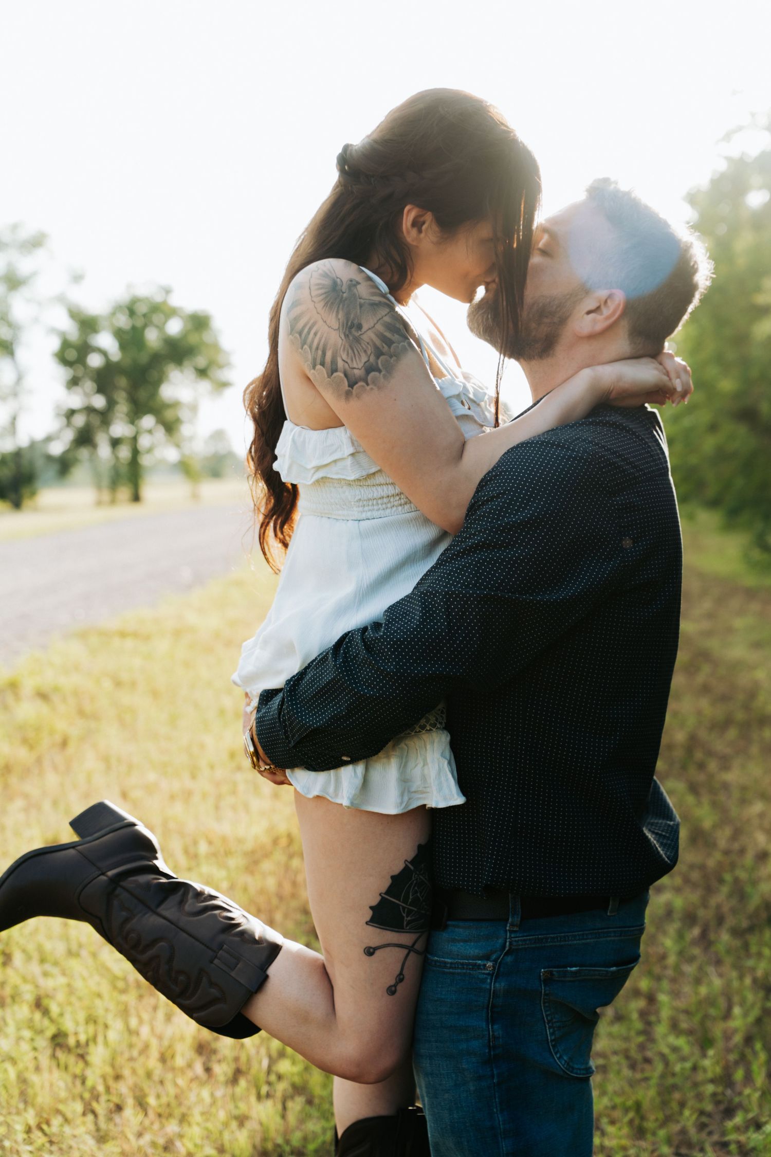 A couple shares a romantic kiss outdoors, man lifting woman in white dress with tattoos, backlit by golden sunlight.
