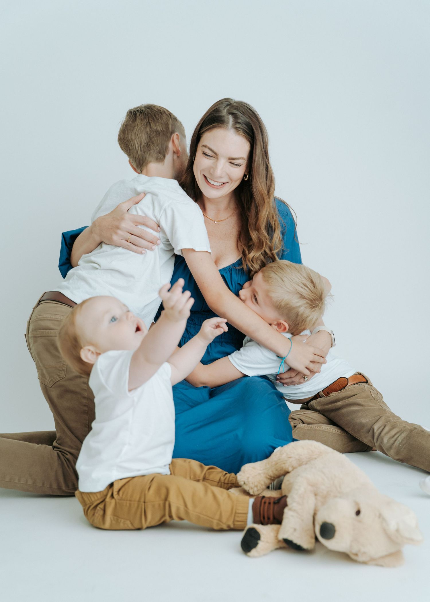A smiling brunette mother in a blue dress cuddles her three young sons on a white studio floor.