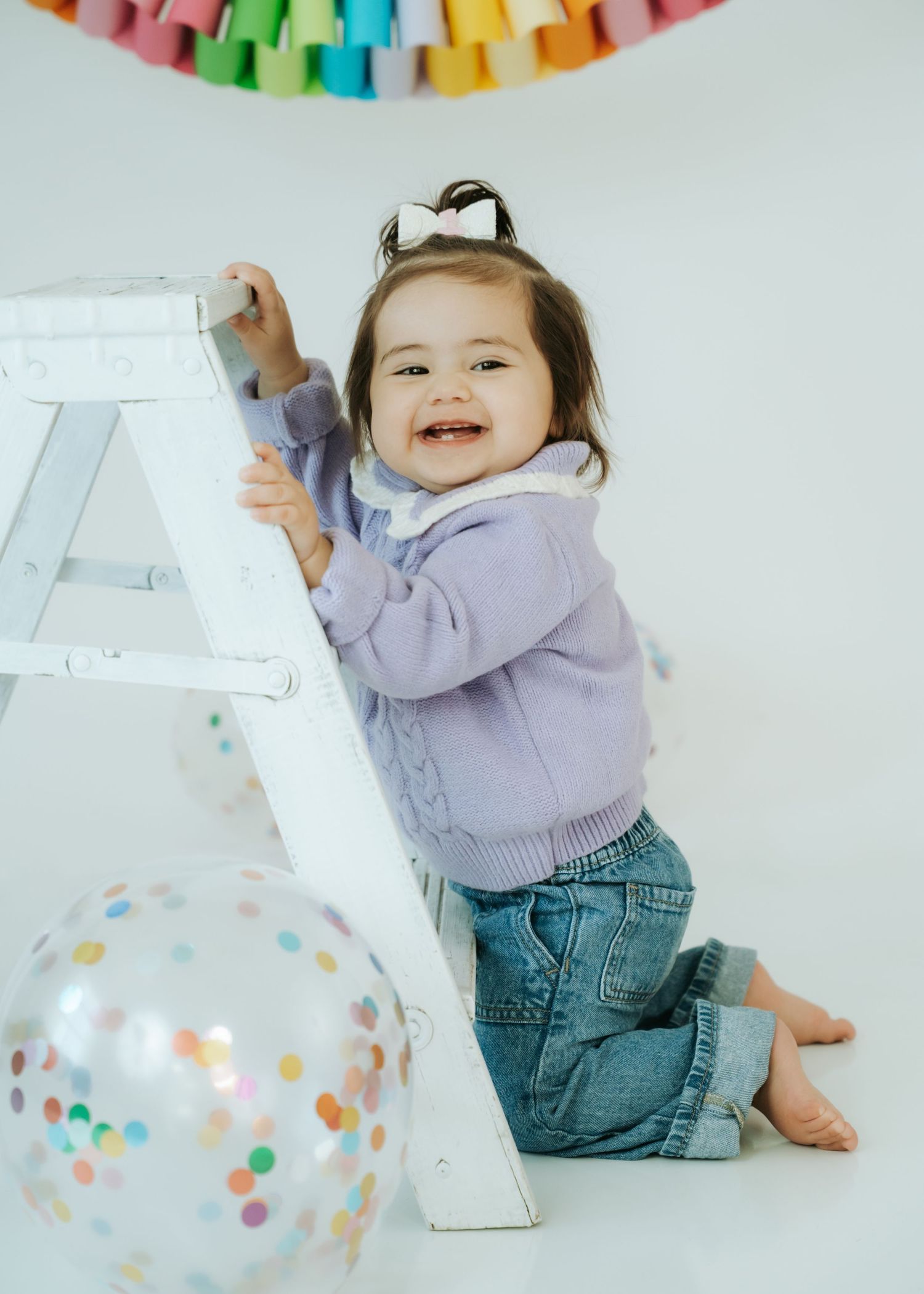 Happy toddler girl in purple sweater and jeans smiles while holding a white ladder near balloons.