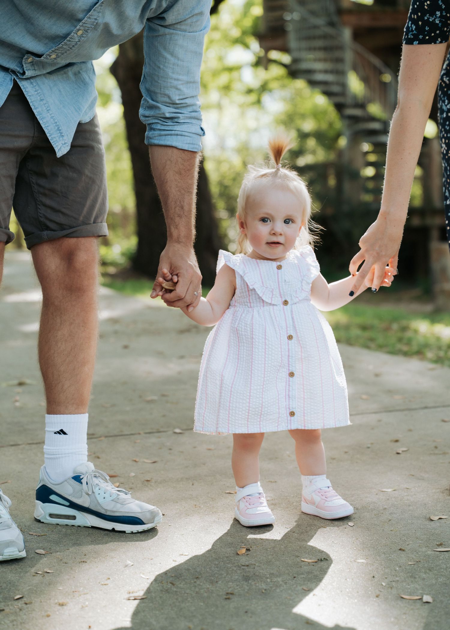 Toddler girl in white dress holds parents hands while walking on sunny outdoor path.