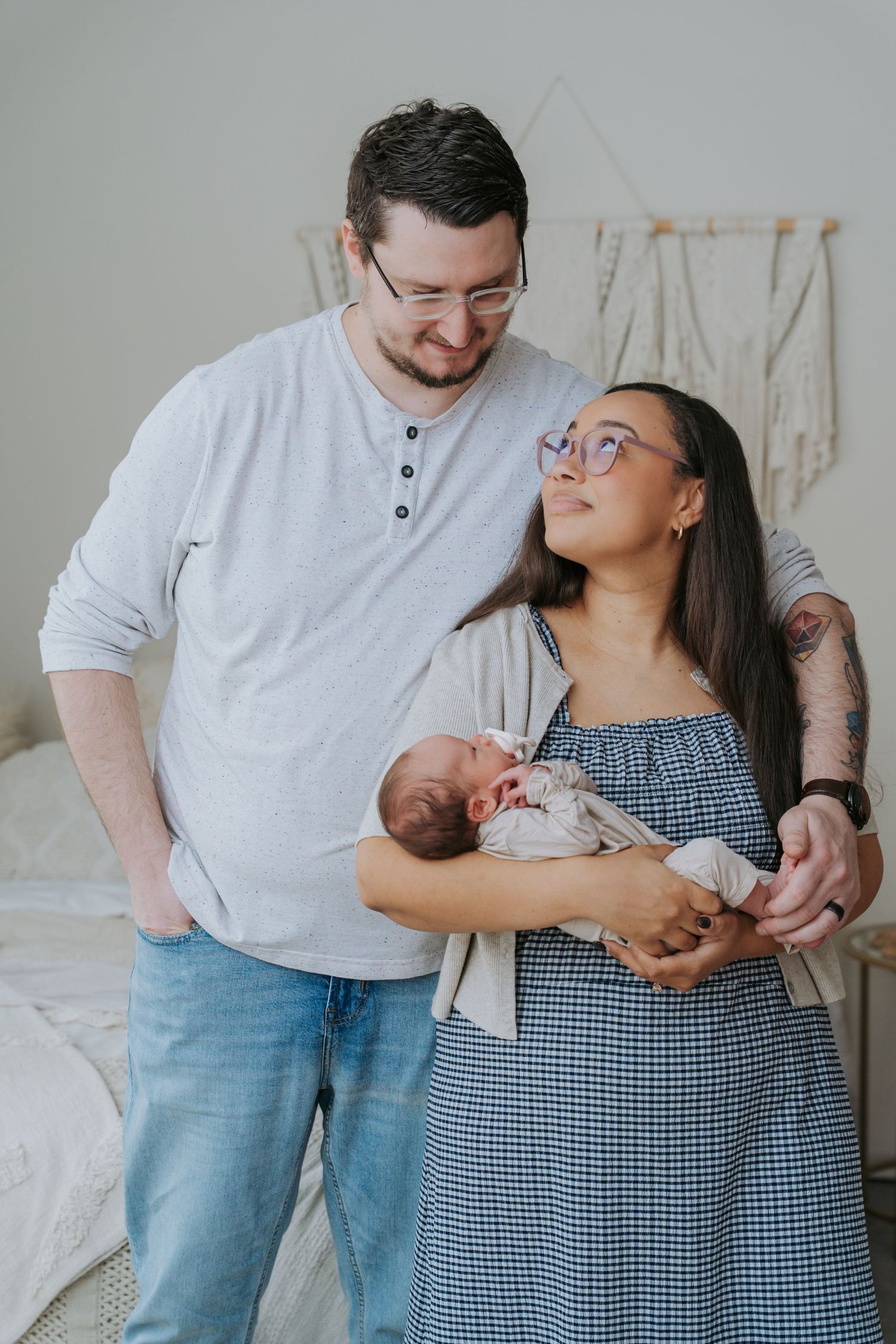 New parents smile at newborn baby in cozy bedroom, mother in blue dress and father in gray shirt with glasses.