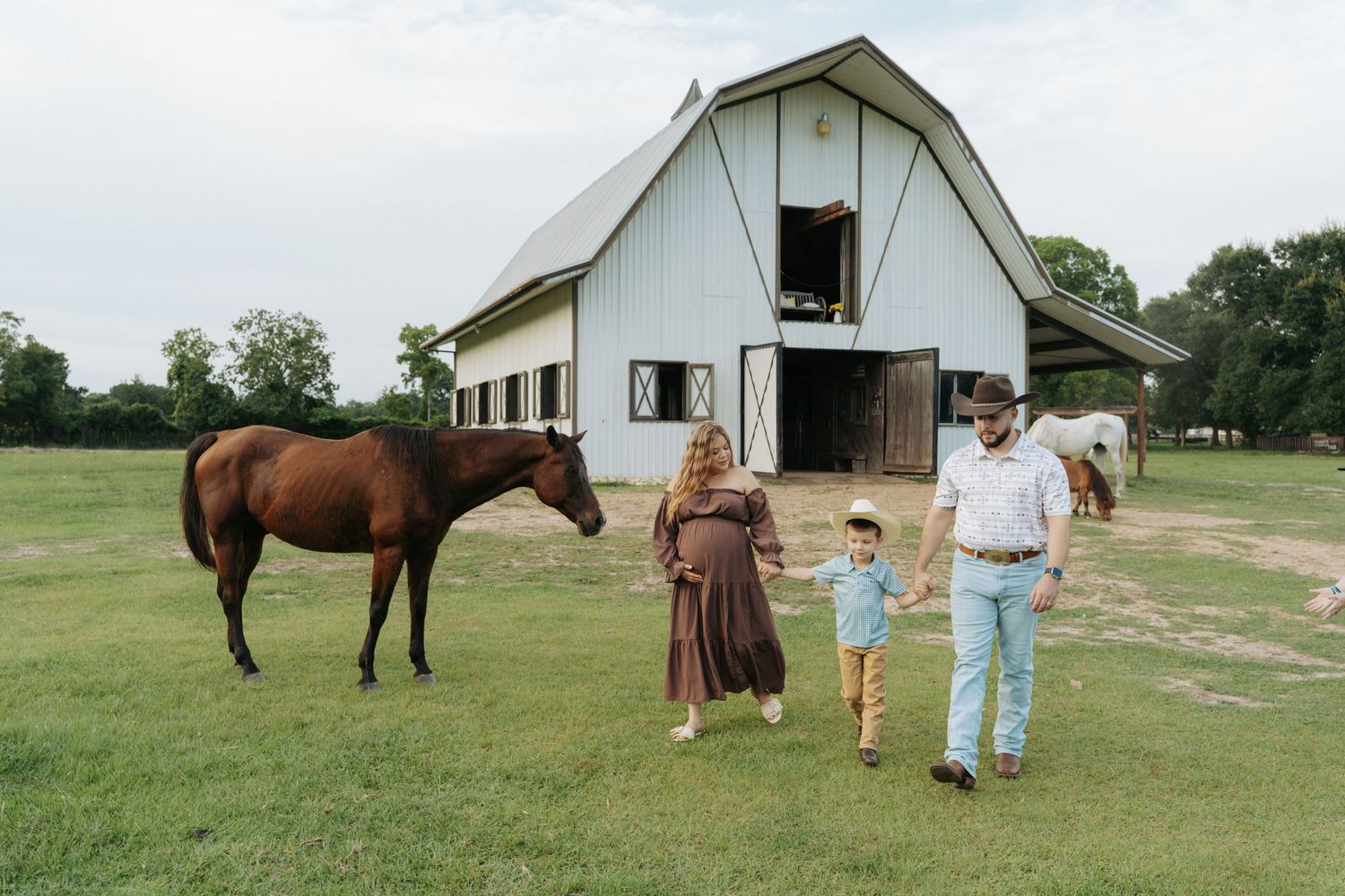 A family walks near a brown horse by a white barn on a green farm, with the mother visibly pregnant.