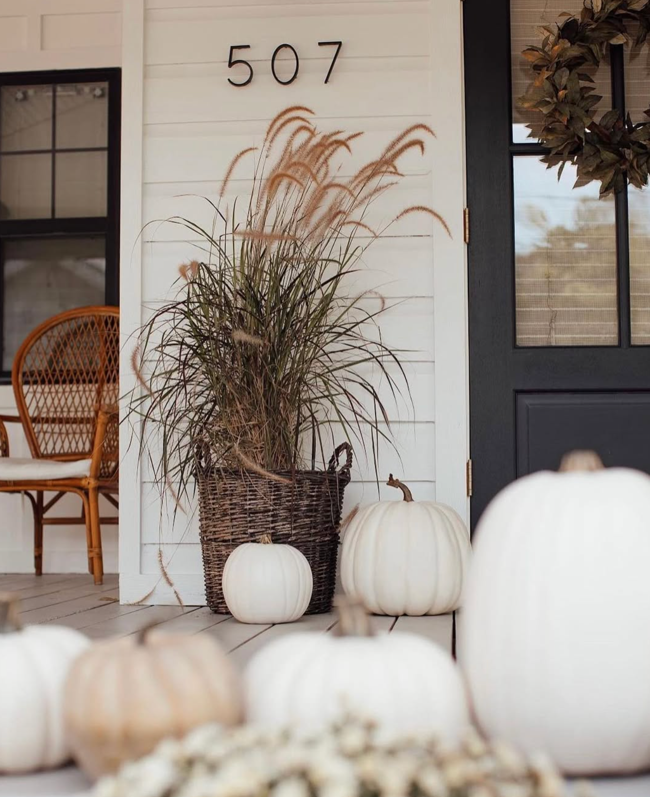 White pumpkins and ornamental grass in wicker basket decorate a farmhouse porch with number 507 and dark front door.