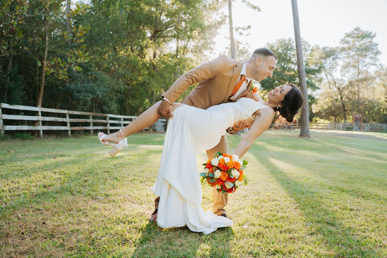 Groom dips bride in white dress holding orange bouquet on green lawn with white fence and trees.