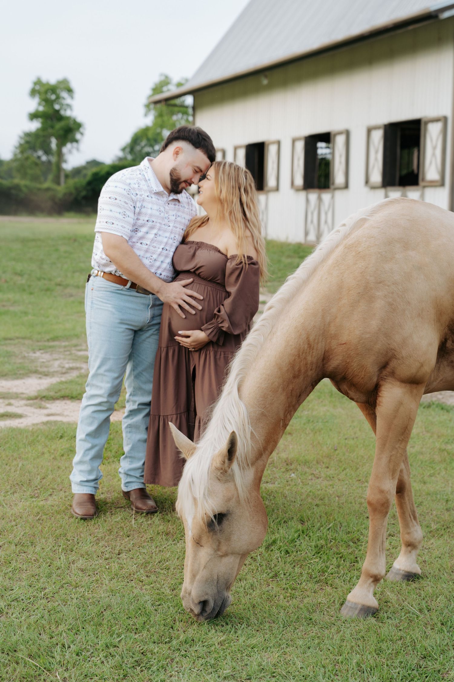 Pregnant couple embracing near a white barn while a palomino horse grazes on green grass nearby.