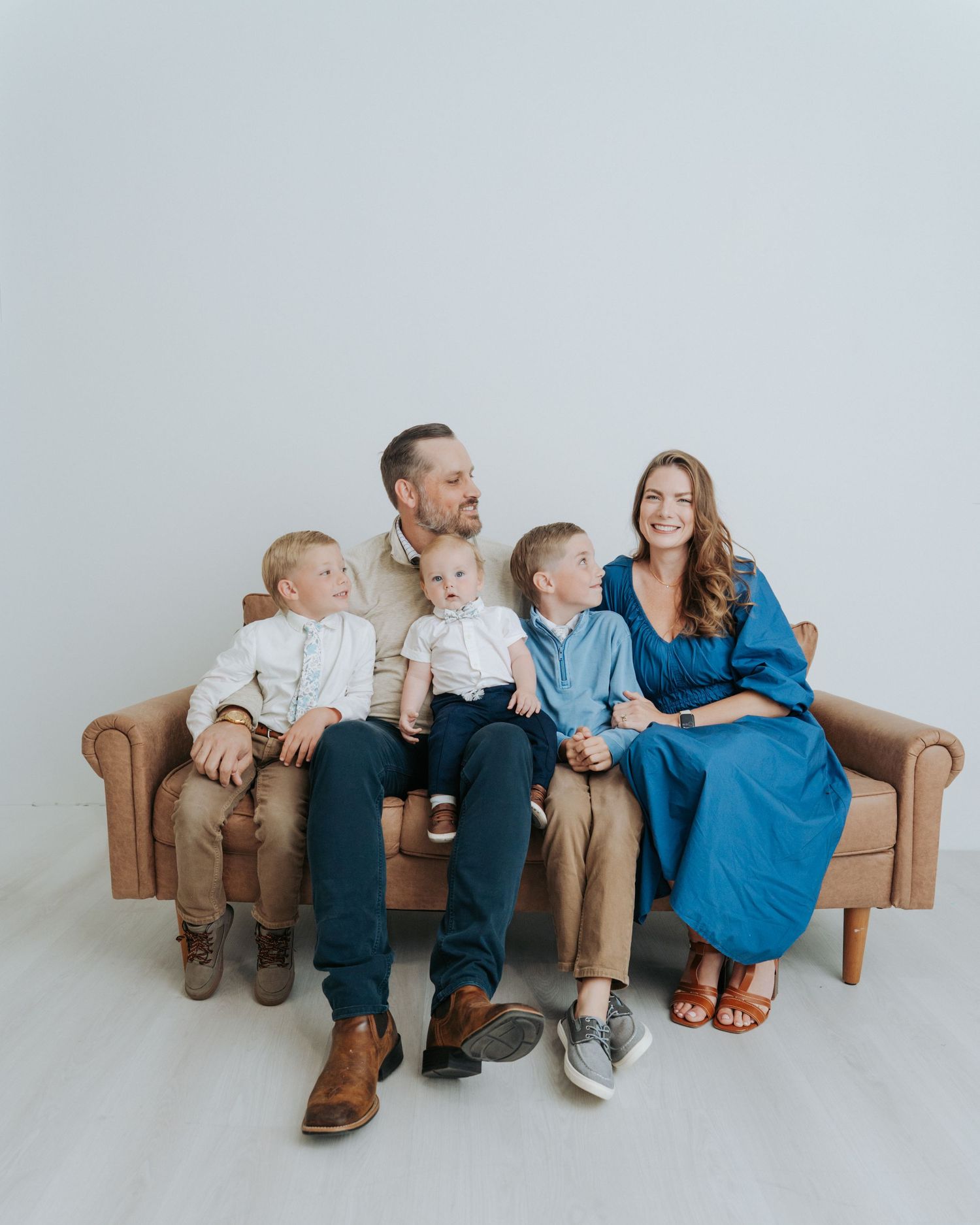 Family of five poses on a brown leather couch in a bright studio, parents with three young boys in coordinated outfits.