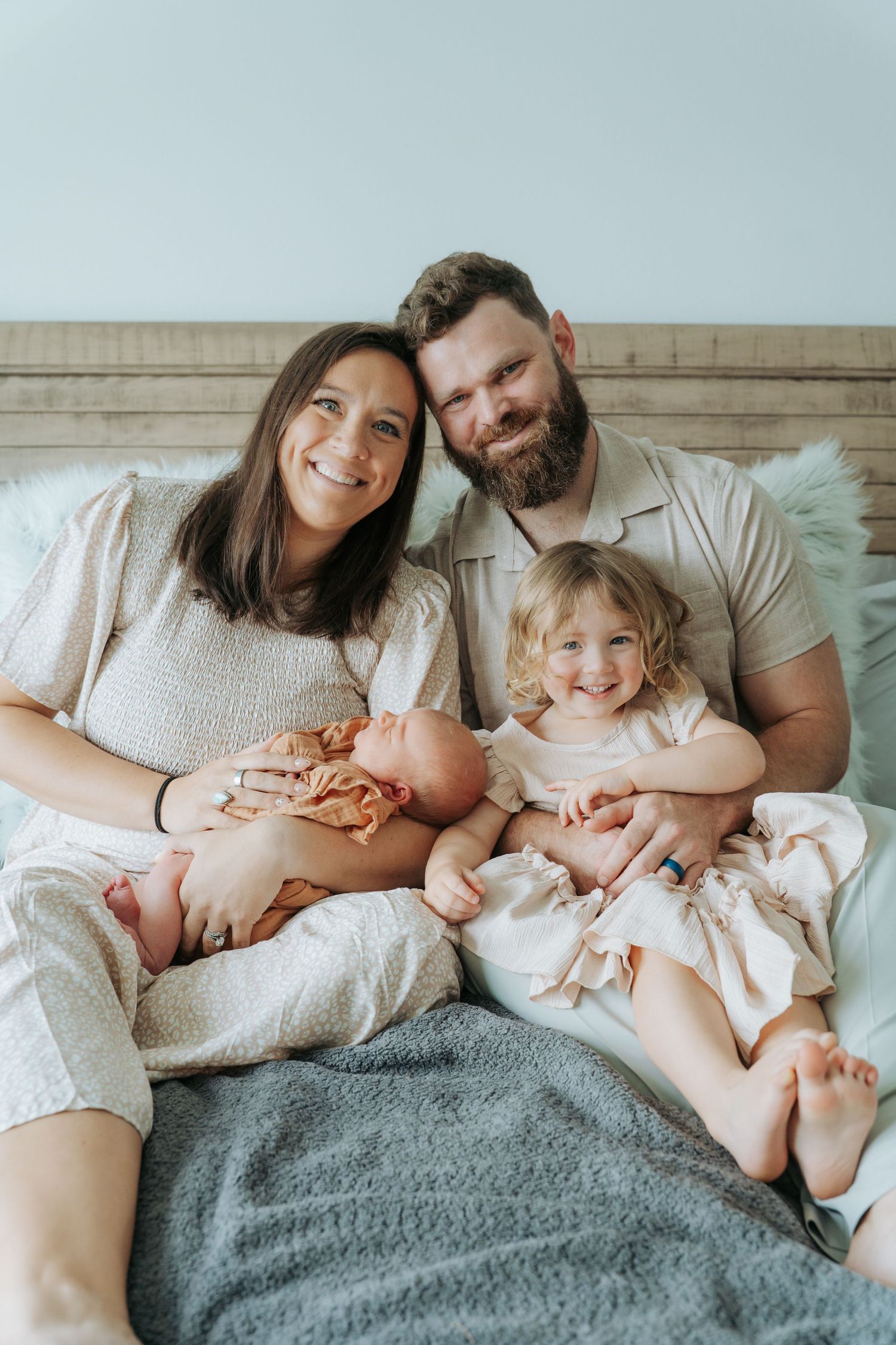 Happy family of four with newborn baby and toddler girl sitting together on a bed, smiling warmly.