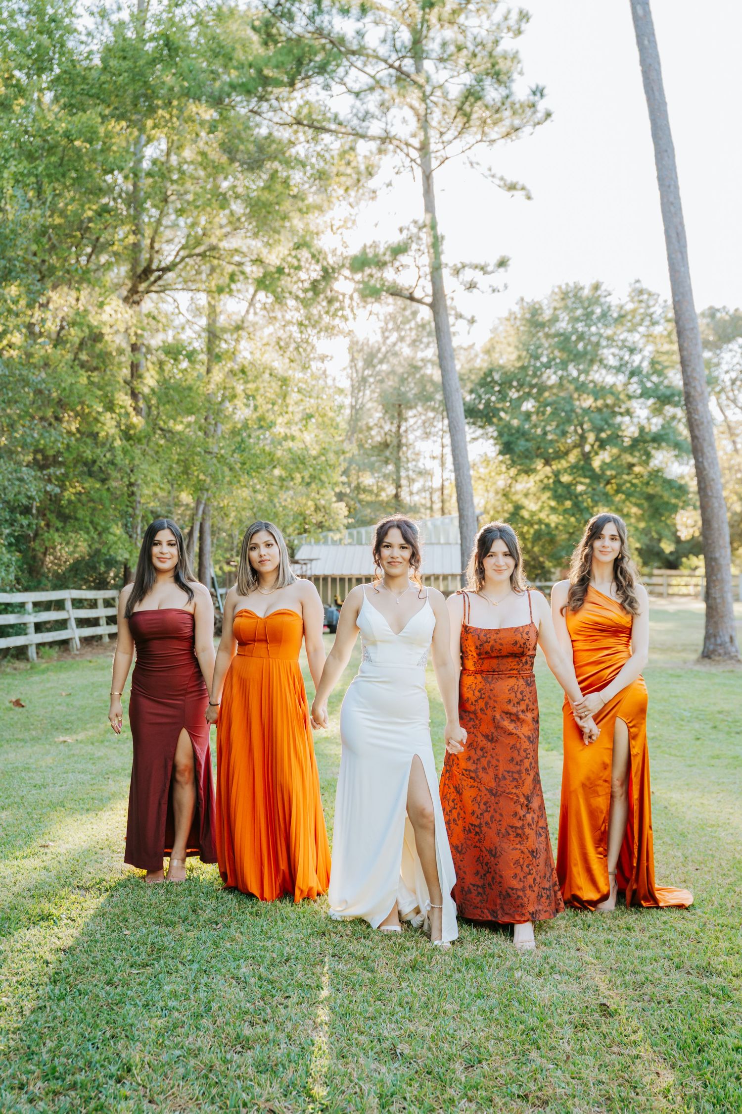 Bride in white gown poses with five bridesmaids in orange and burgundy dresses outdoors on green lawn.