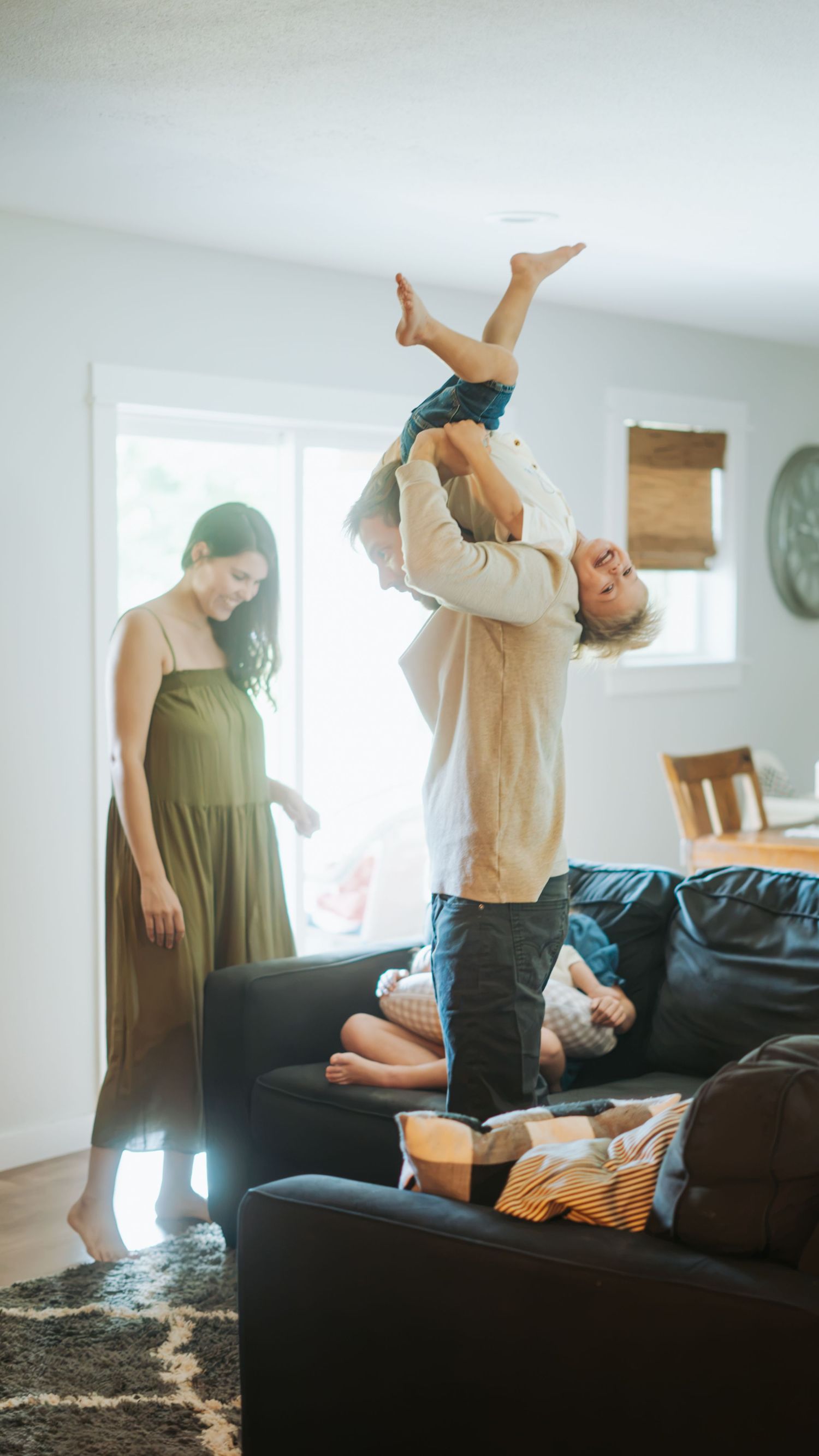 A father joyfully lifts a child overhead in a bright living room while a woman in a green dress watches and another child relaxes on a sofa.