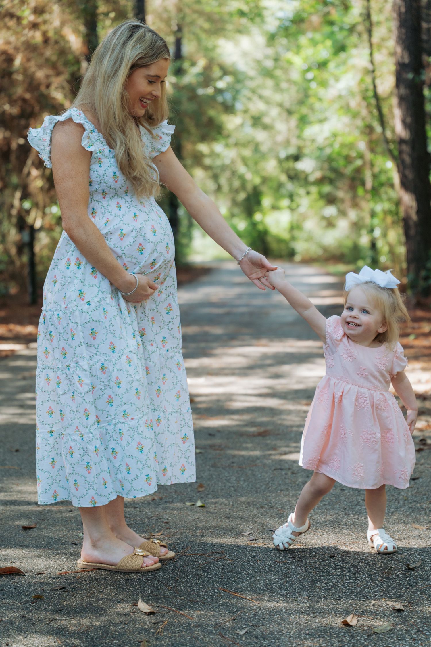 Pregnant woman in floral dress holds hands with toddler girl in pink dress on a tree-lined path.