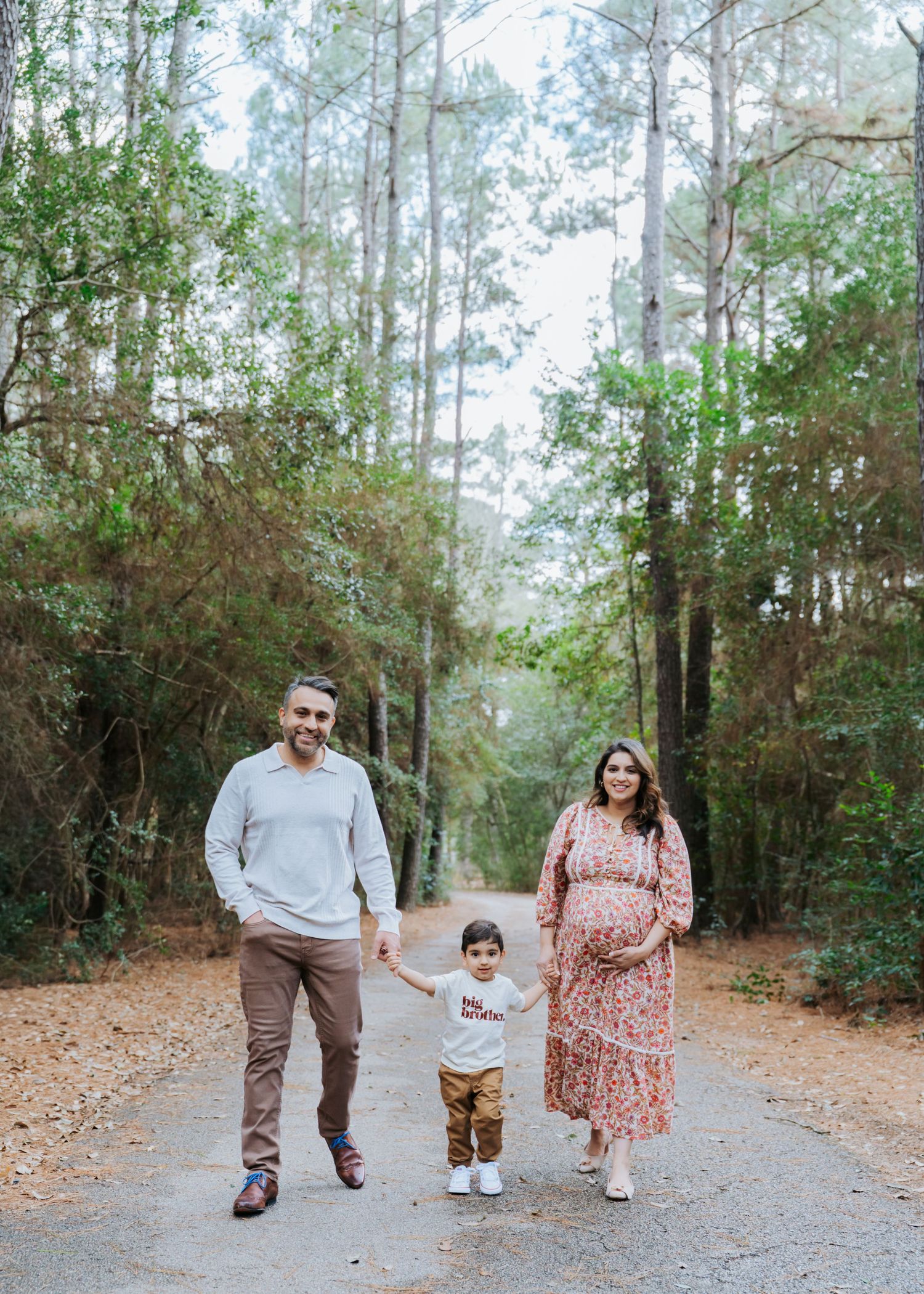 Expecting family of three walks down a pine forest path, mother in floral maternity dress holding toddlers hand.