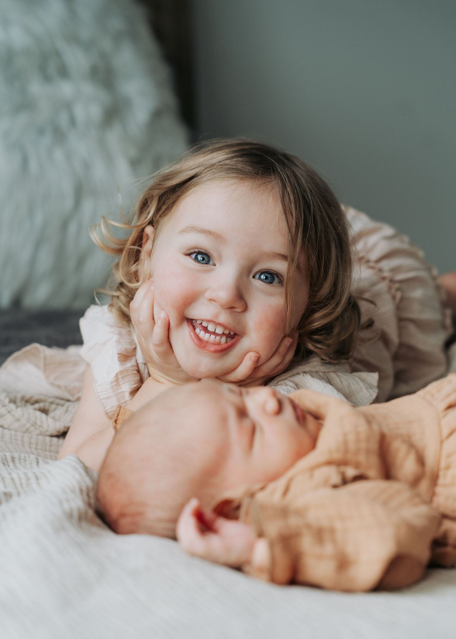 Smiling toddler girl with blue eyes resting chin on hand next to sleeping newborn sibling.