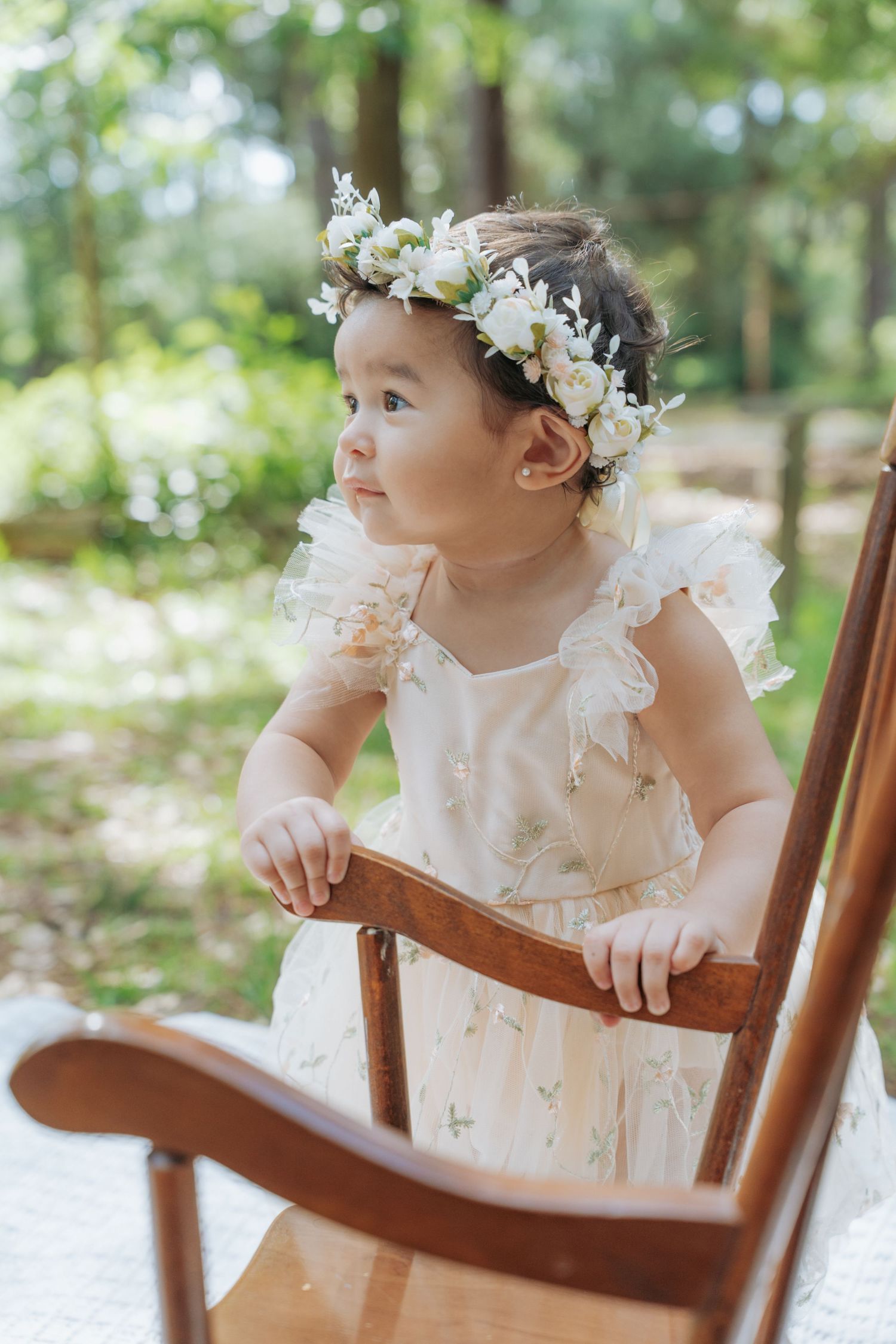 Toddler girl in floral crown and white dress standing by wooden rocking chair in sunny garden.
