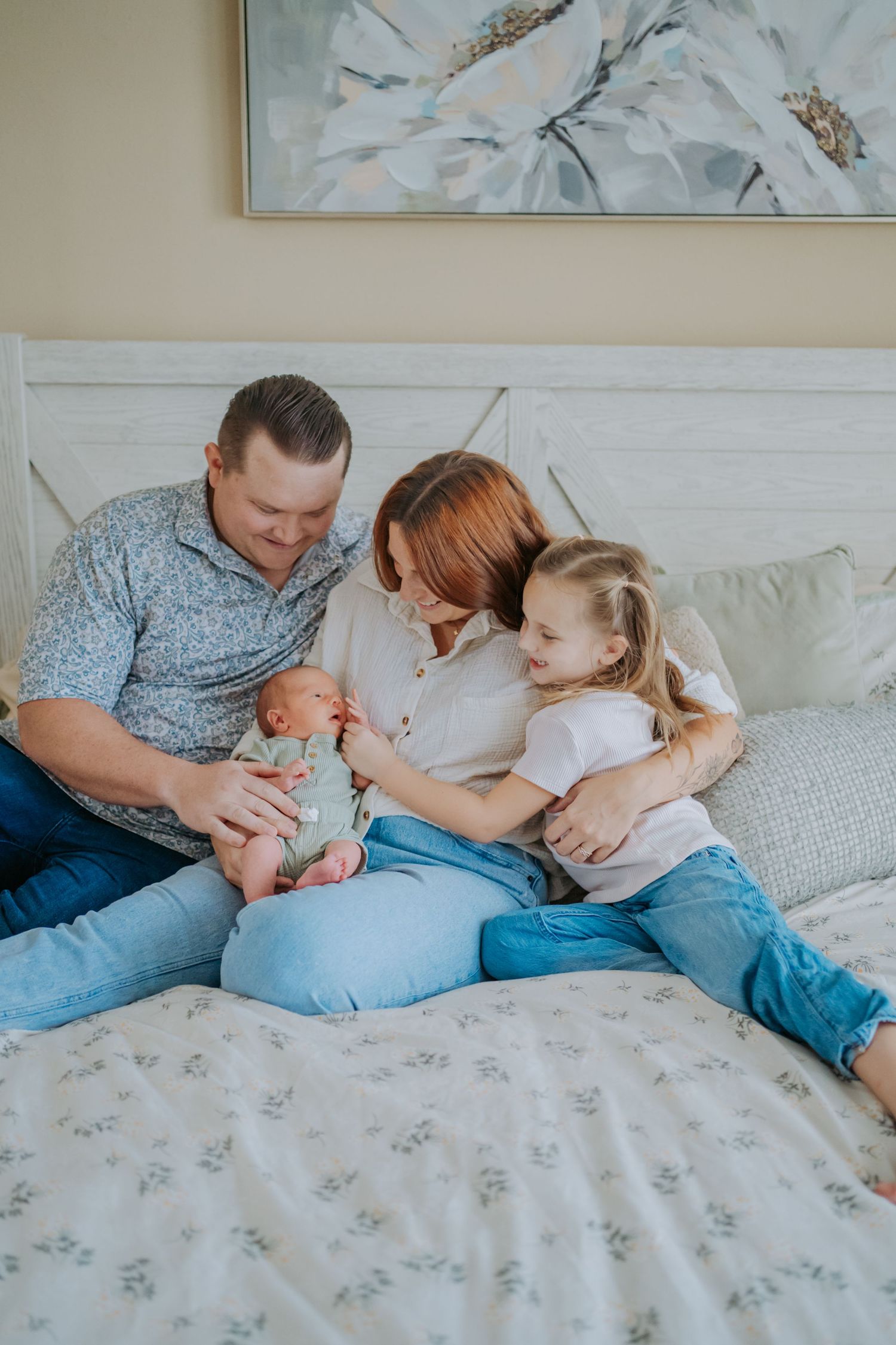Family of four sitting on bed, parents admiring newborn baby while young daughter smiles and hugs mom.
