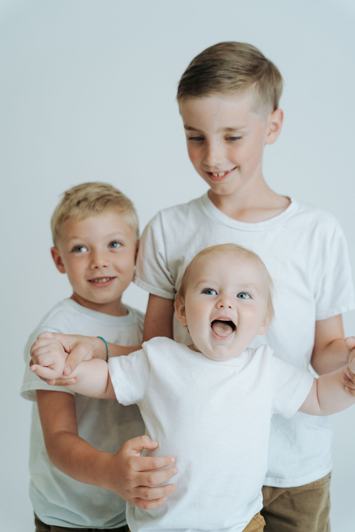 Three brothers in white shirts smiling and hugging together, including a laughing baby, in a studio photo.