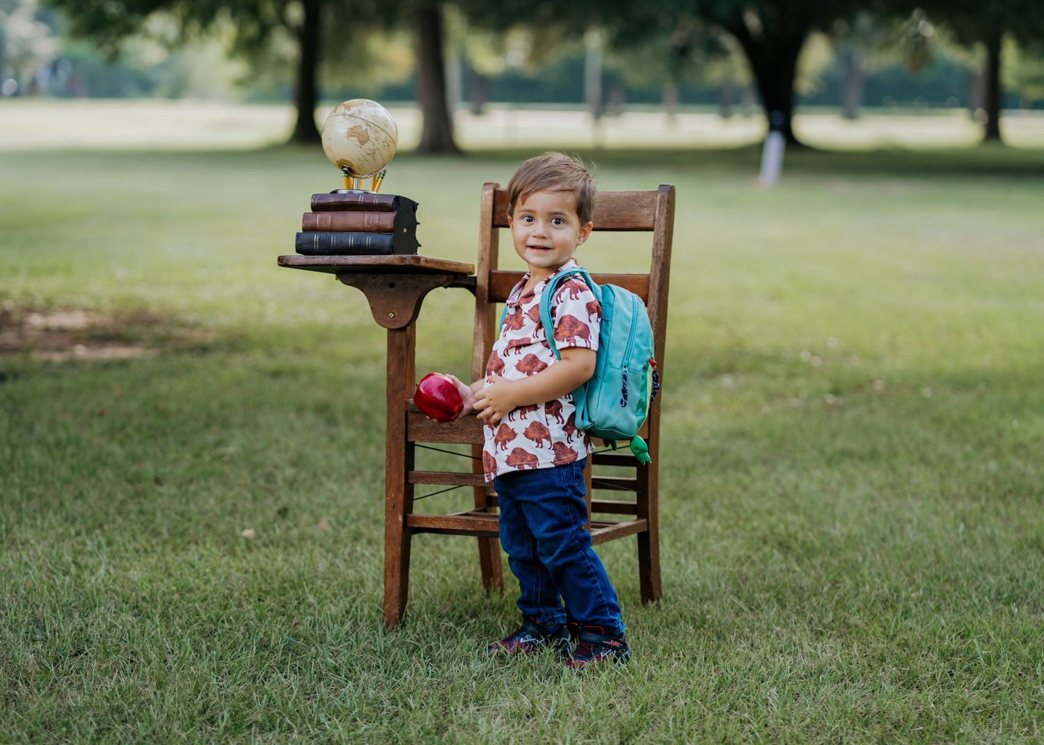 Young boy with backpack stands beside vintage wooden desk with books outdoors on green grass for back-to-school photo.