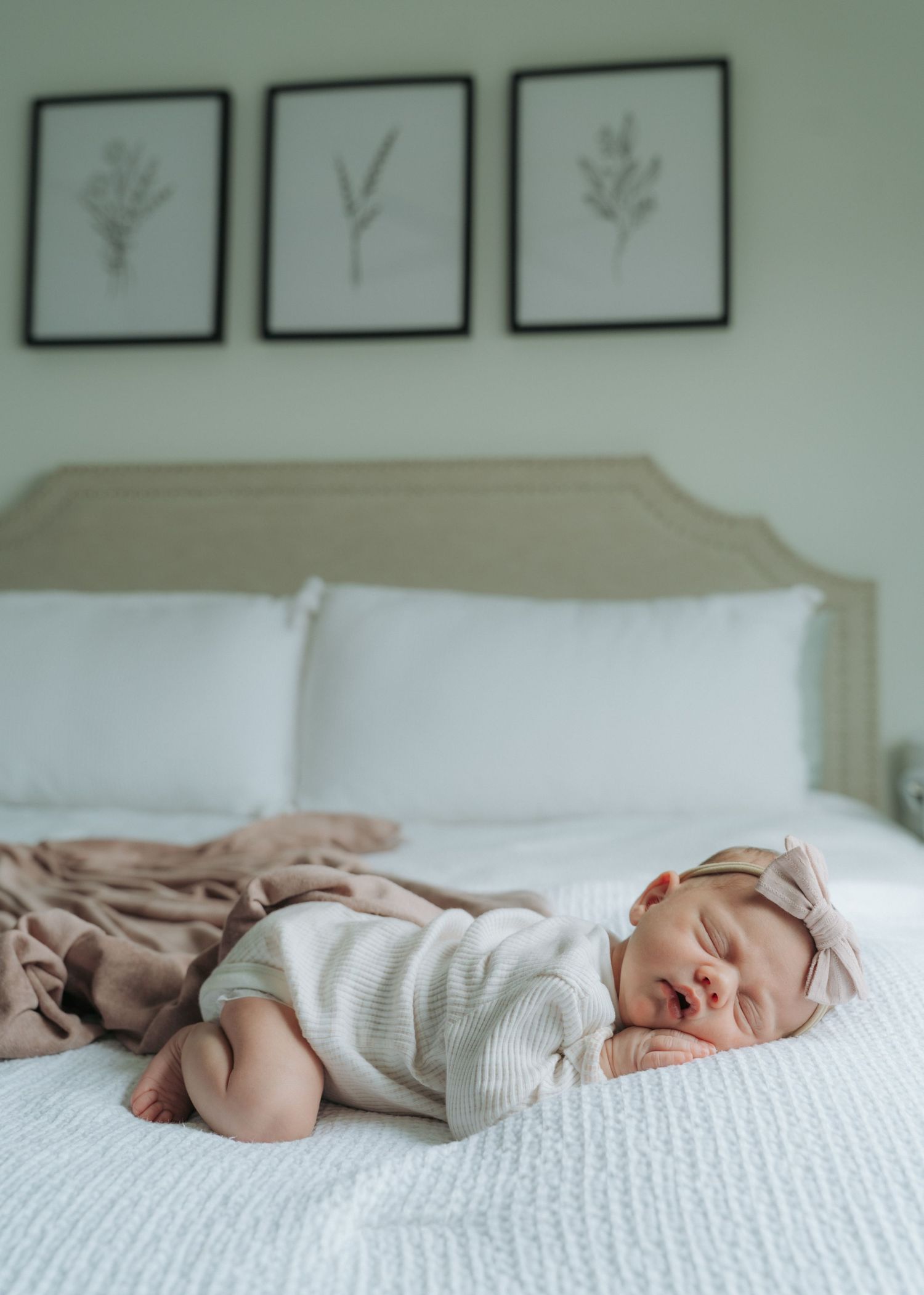 Peaceful newborn baby girl sleeping on white bed wearing striped onesie and pink bow headband.