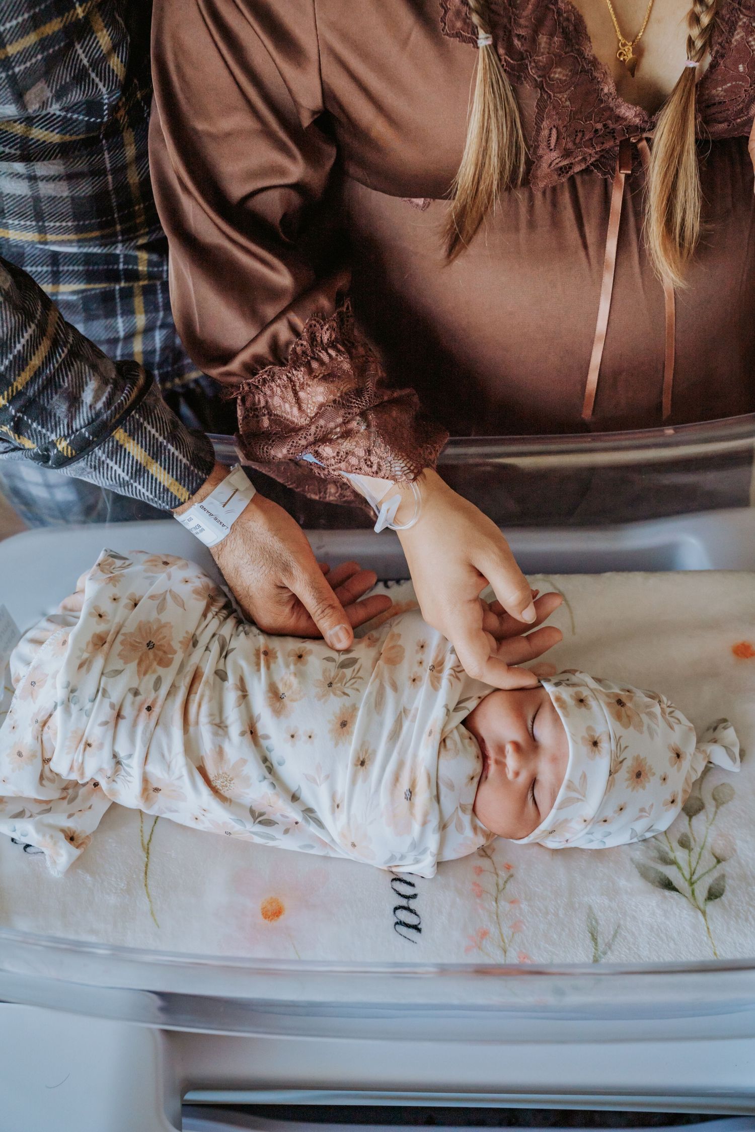 New parents gently touch their swaddled newborn baby in a floral blanket in the hospital bassinet.