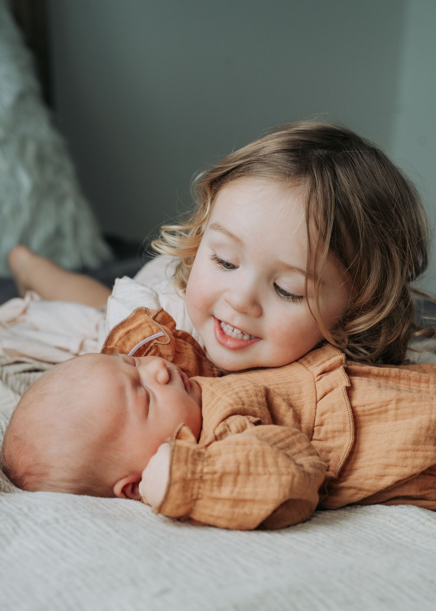Adorable toddler girl smiles while snuggling close to her sleeping newborn sibling on a soft bed.
