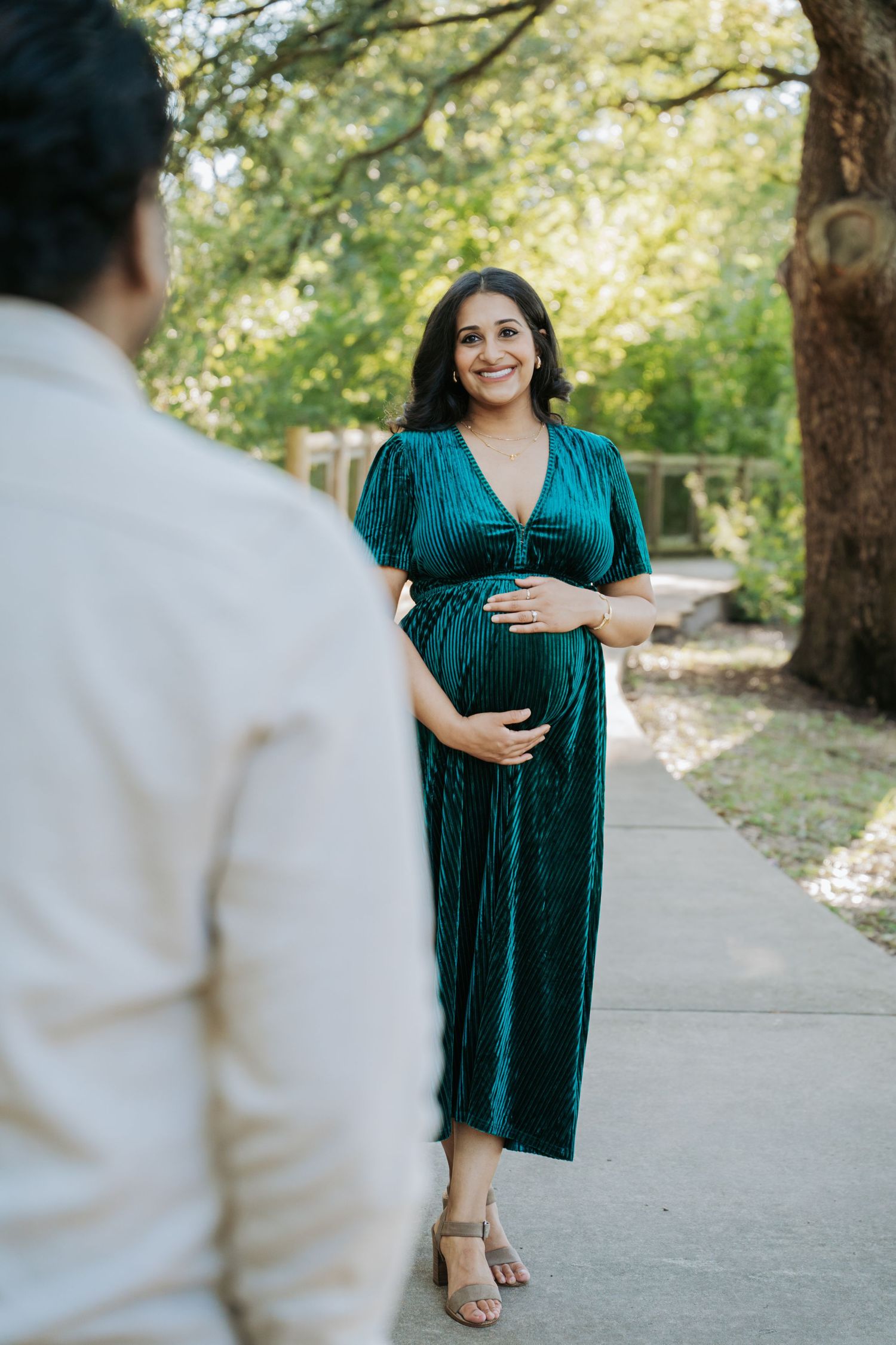 Pregnant woman in emerald green velvet dress cradles baby bump on a tree-lined outdoor walkway.