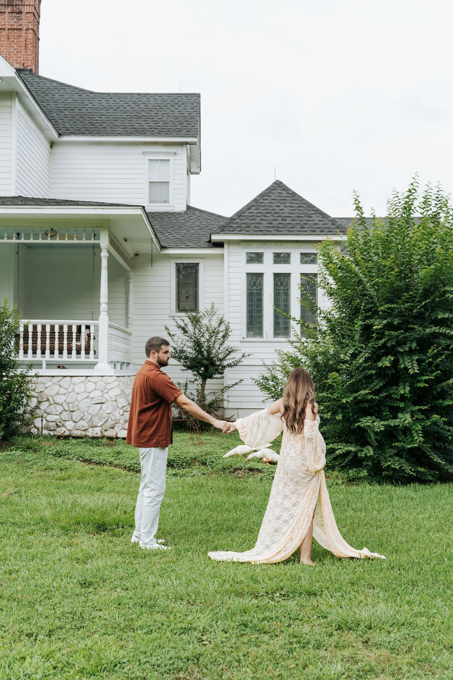 Couple holding hands and dancing on green lawn in front of a white farmhouse with a porch and lush shrubs.