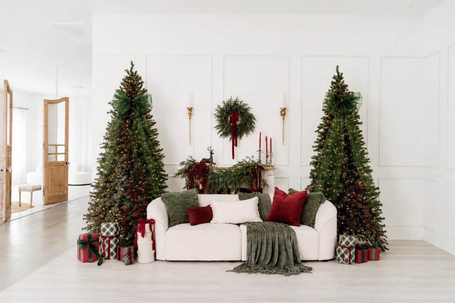 Festive living room with two Christmas trees, white sofa, red pillows, wrapped gifts, and holiday wreath on white wall.