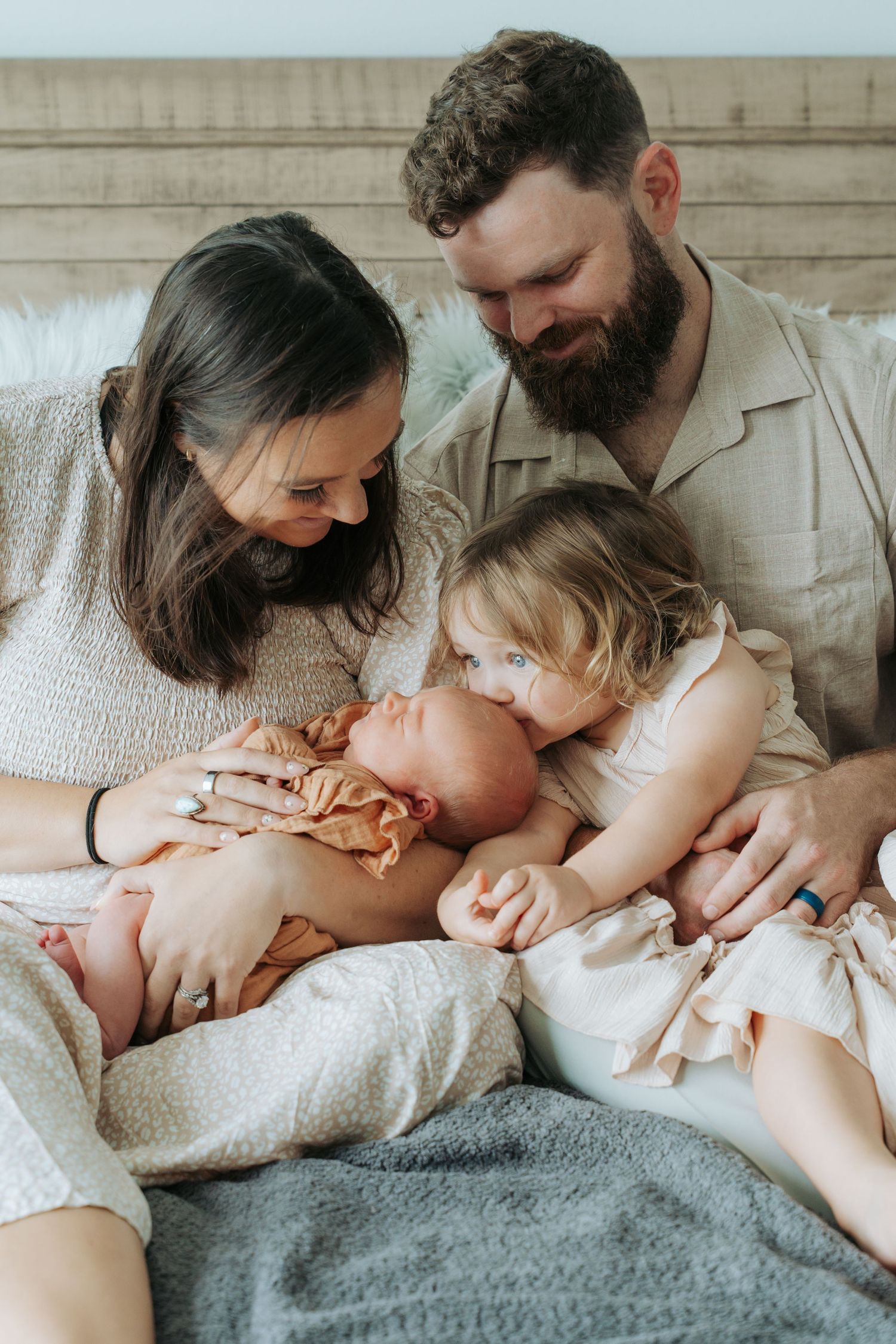 A loving family of four cuddles on a bed with a newborn wrapped in an orange swaddle.