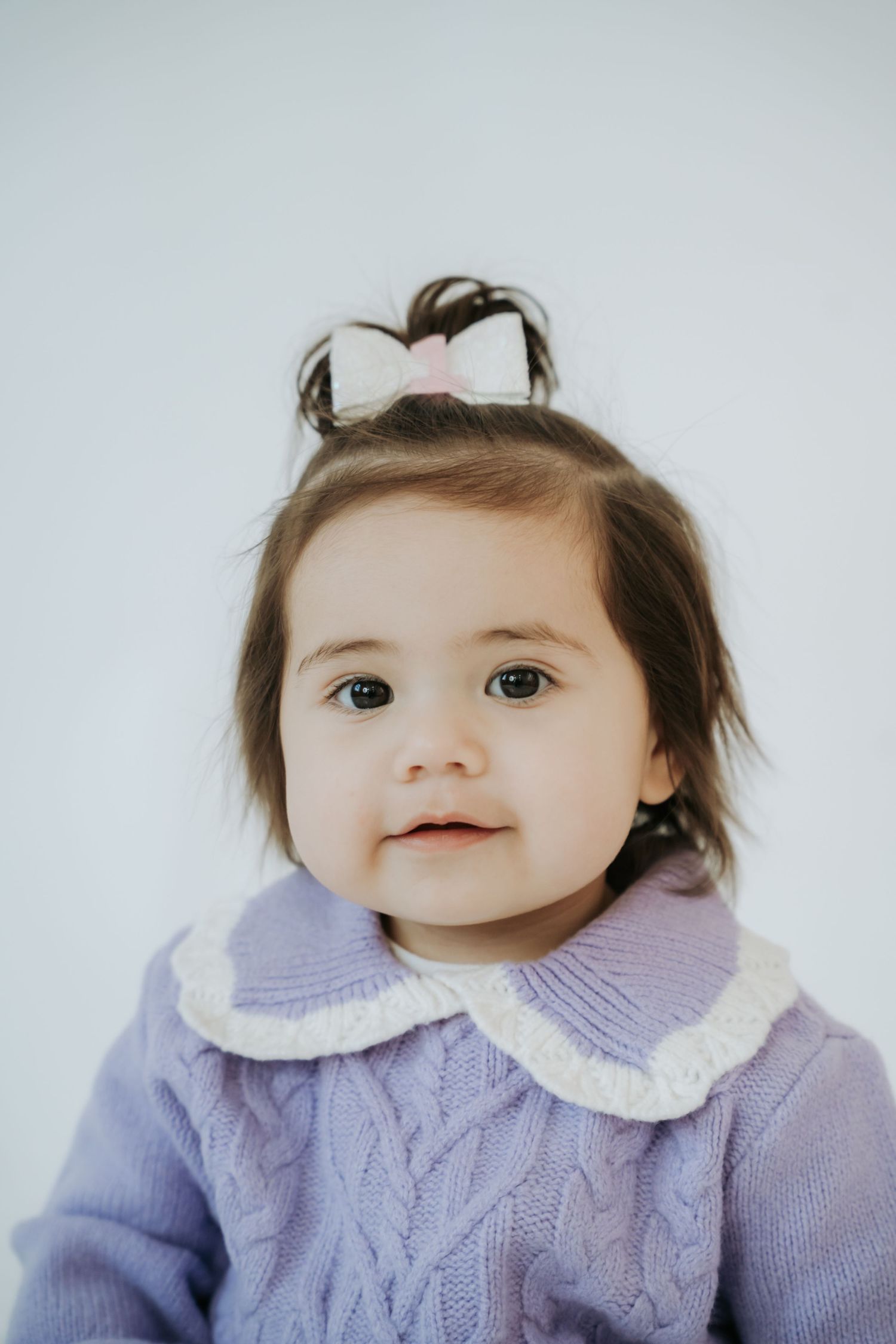 Cute baby girl in a purple cable-knit sweater with white collar poses against a white background.