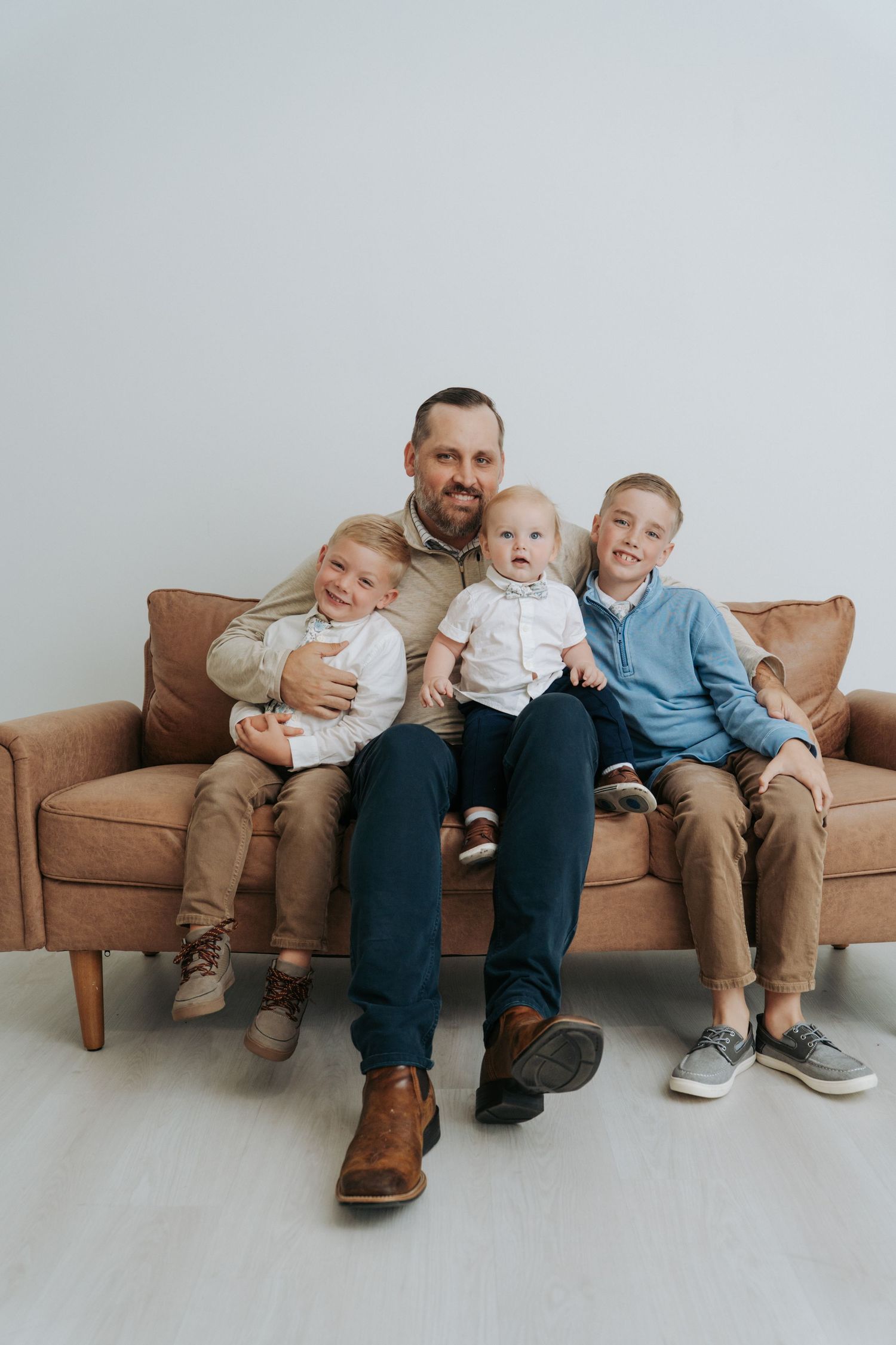 Father sitting on brown leather sofa with three young sons, smiling for a family portrait photo.