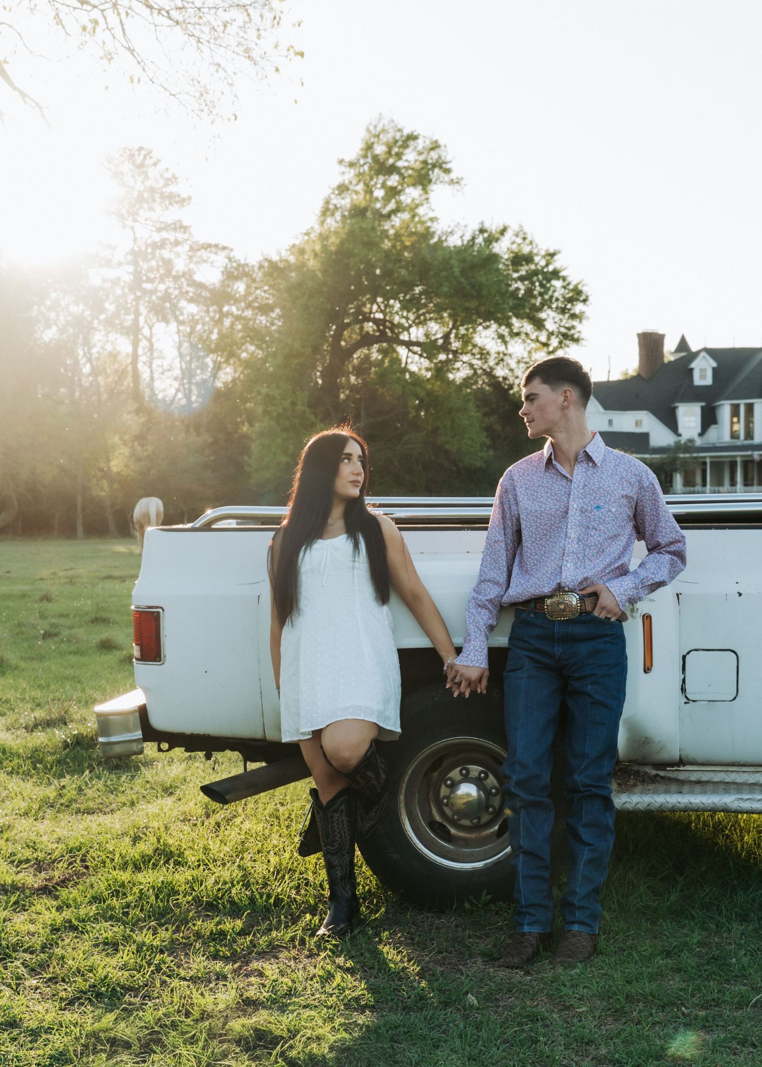 Young couple holds hands leaning against vintage white truck at golden hour on farm.