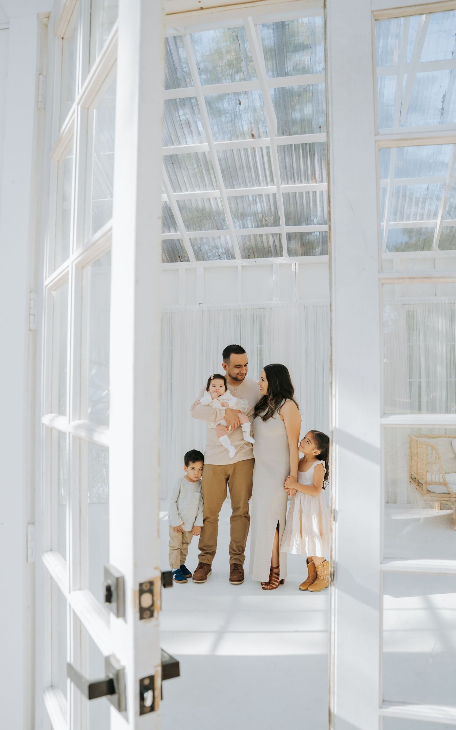 Family of four photographed through white French doors in bright white greenhouse setting.