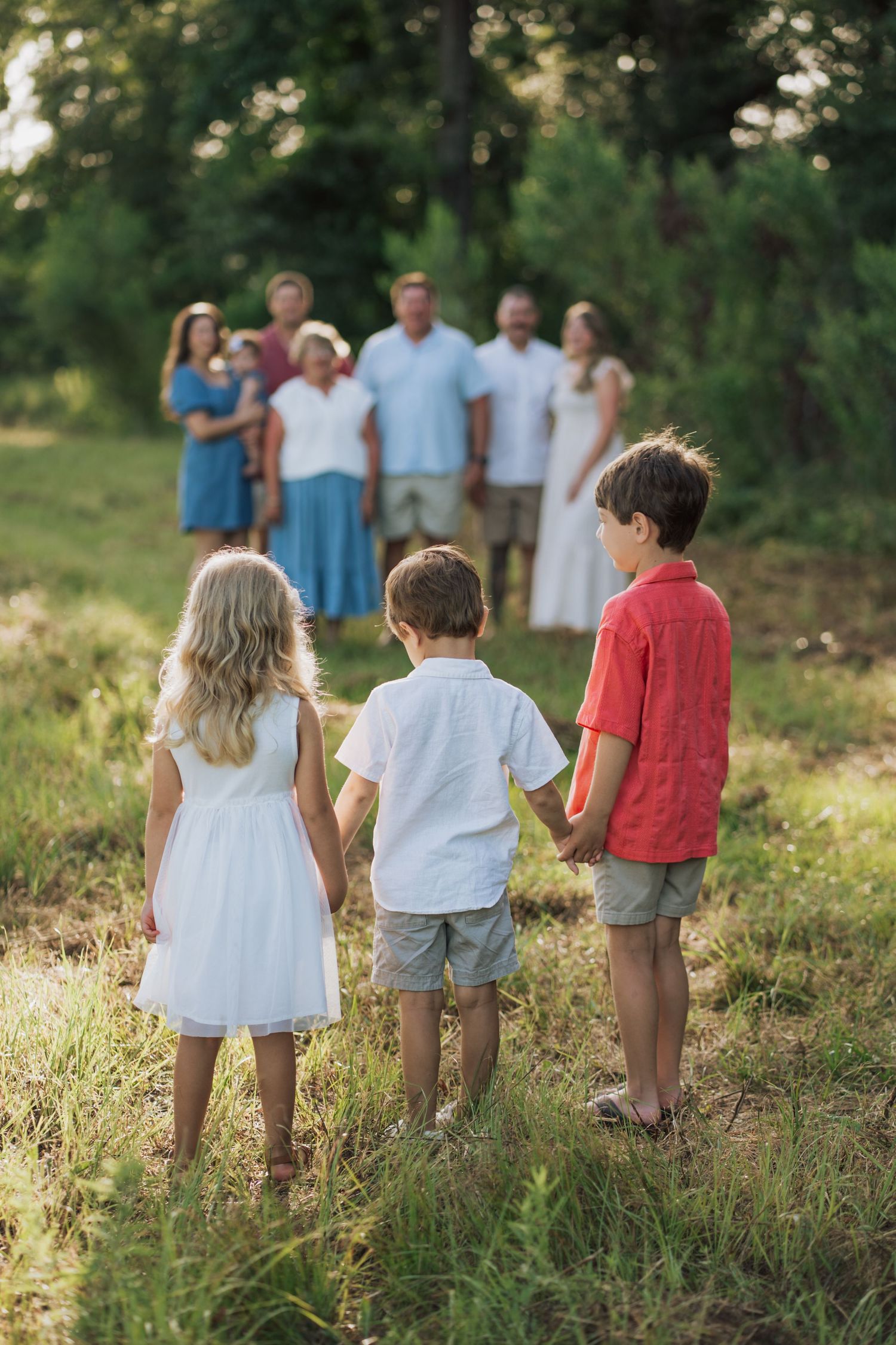 Three children holding hands facing extended family gathered in lush green outdoor field.