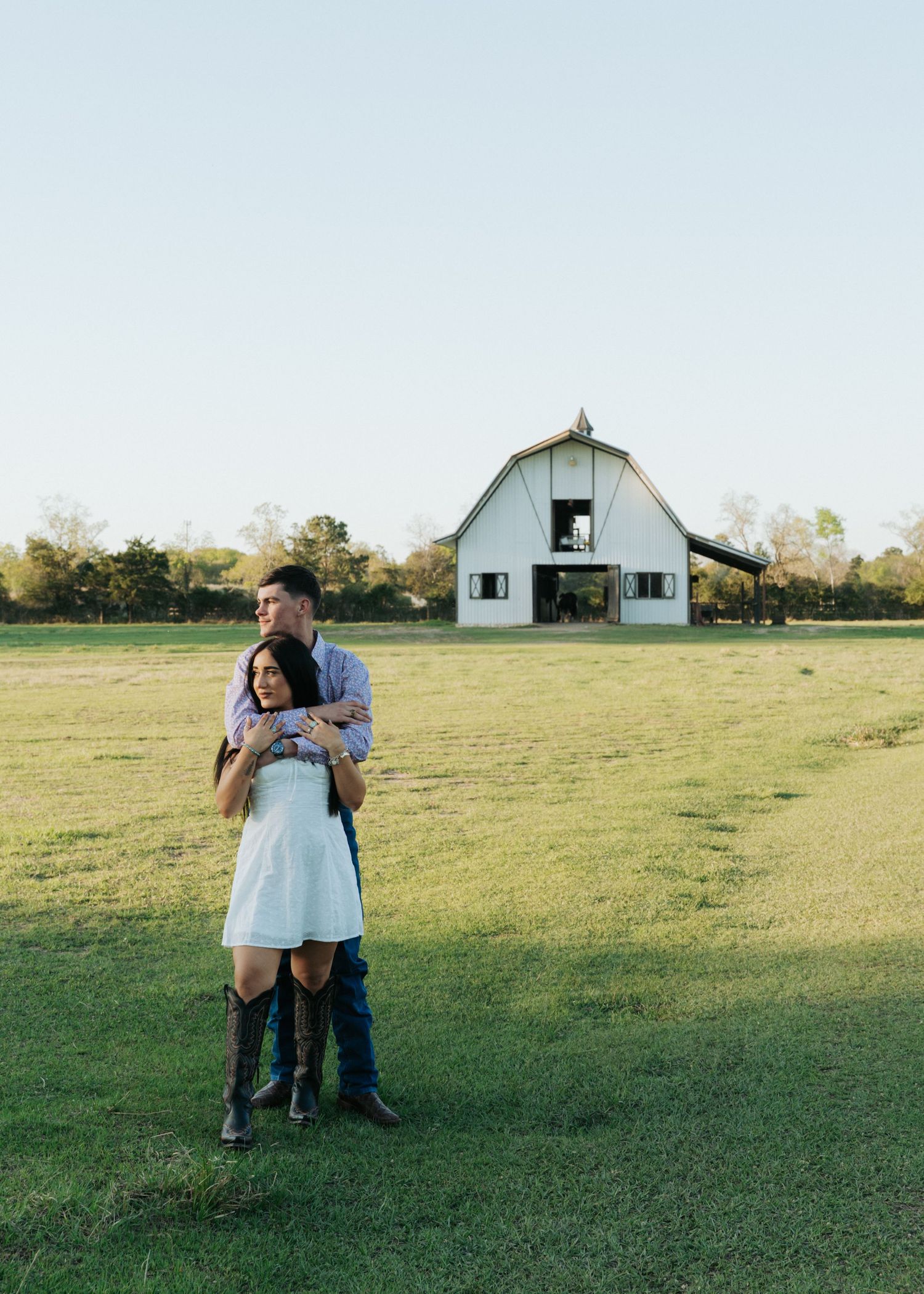 Couple embraces in open green field with white barn backdrop during outdoor engagement session.